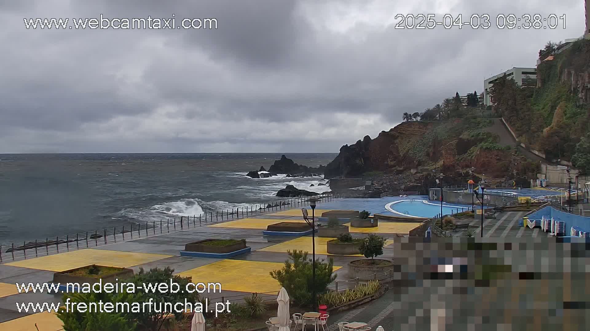 A coastal outdoor scene shows a partly cloudy sky over a rocky shoreline with a swimming pool complex and a choppy ocean.