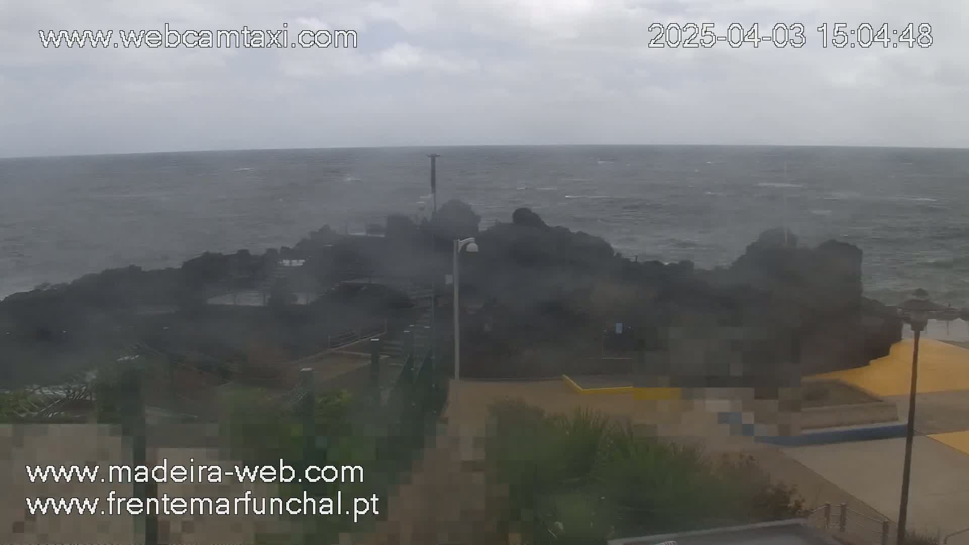 A stormy ocean with choppy waves crashes against dark rocks near a paved area with some vegetation.