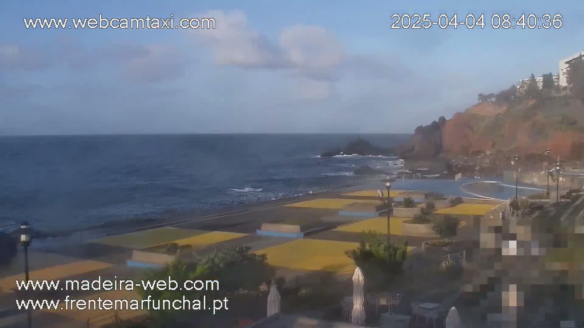 A mostly sunny day shows a view of a rocky coastline with a dark blue ocean, a paved area with yellow and blue sections near a swimming pool, and a reddish-brown cliff in the background.