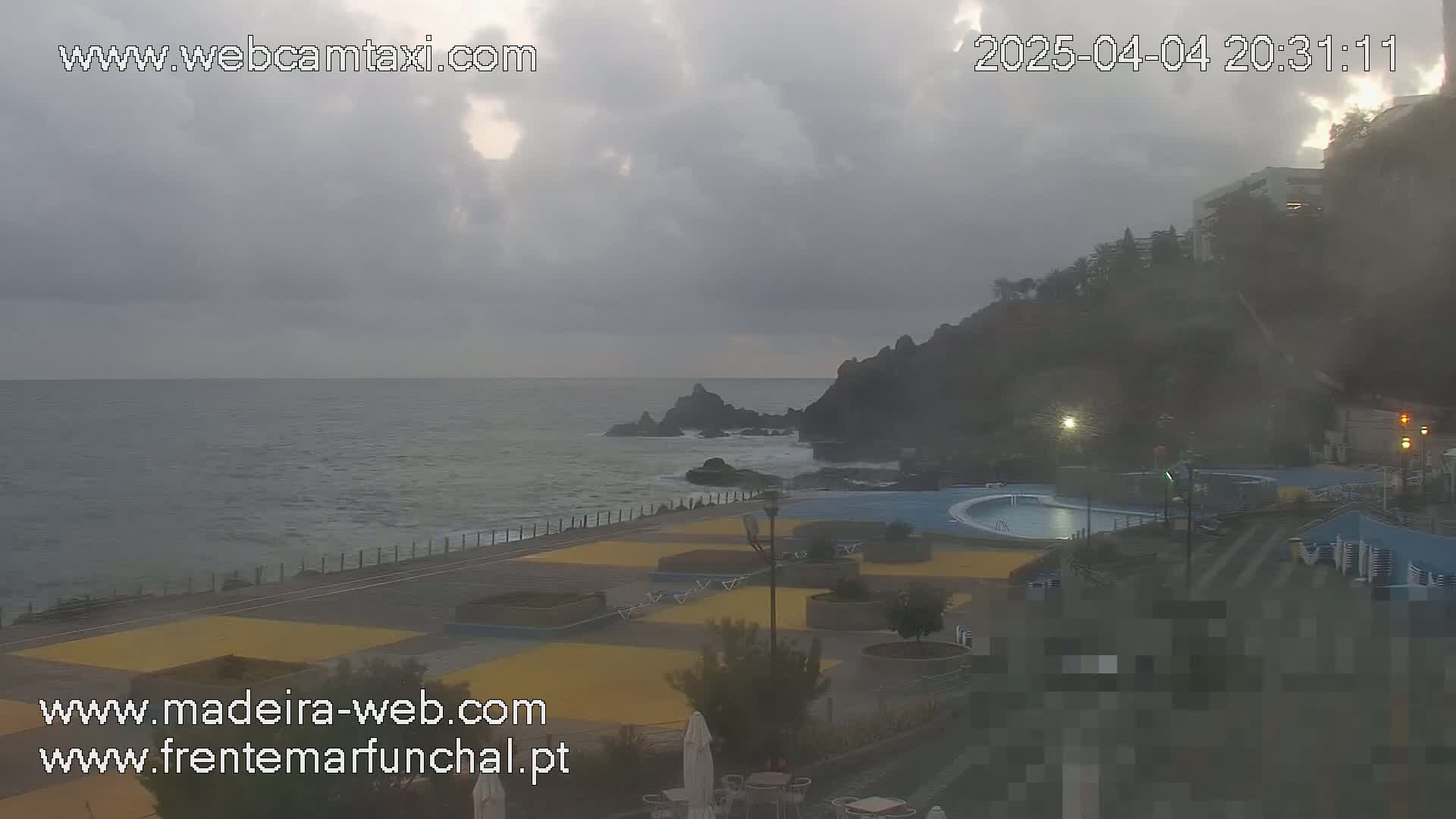 A mostly cloudy, overcast day shows a rocky coastline with an outdoor pool and paved walkway beside a calm ocean.