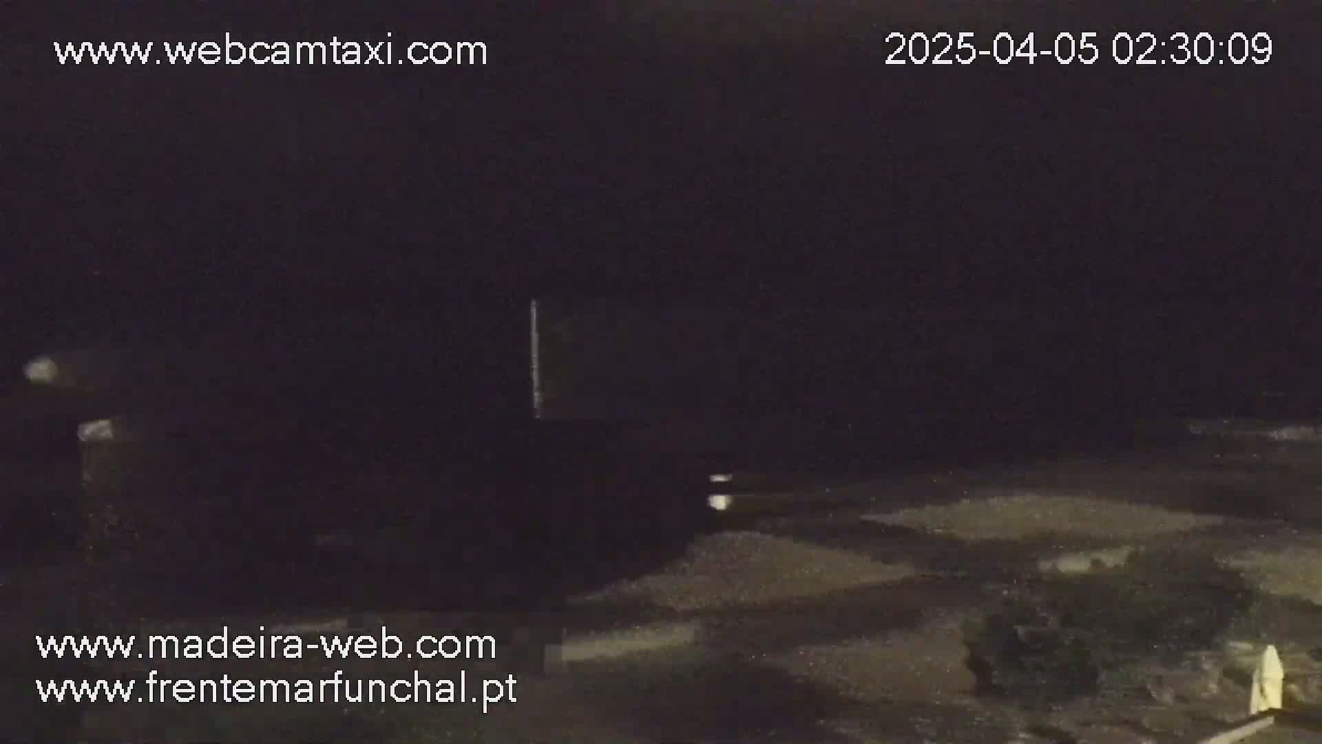 A dark, nighttime scene shows a mostly empty paved area with indistinct objects and some faint lights.
