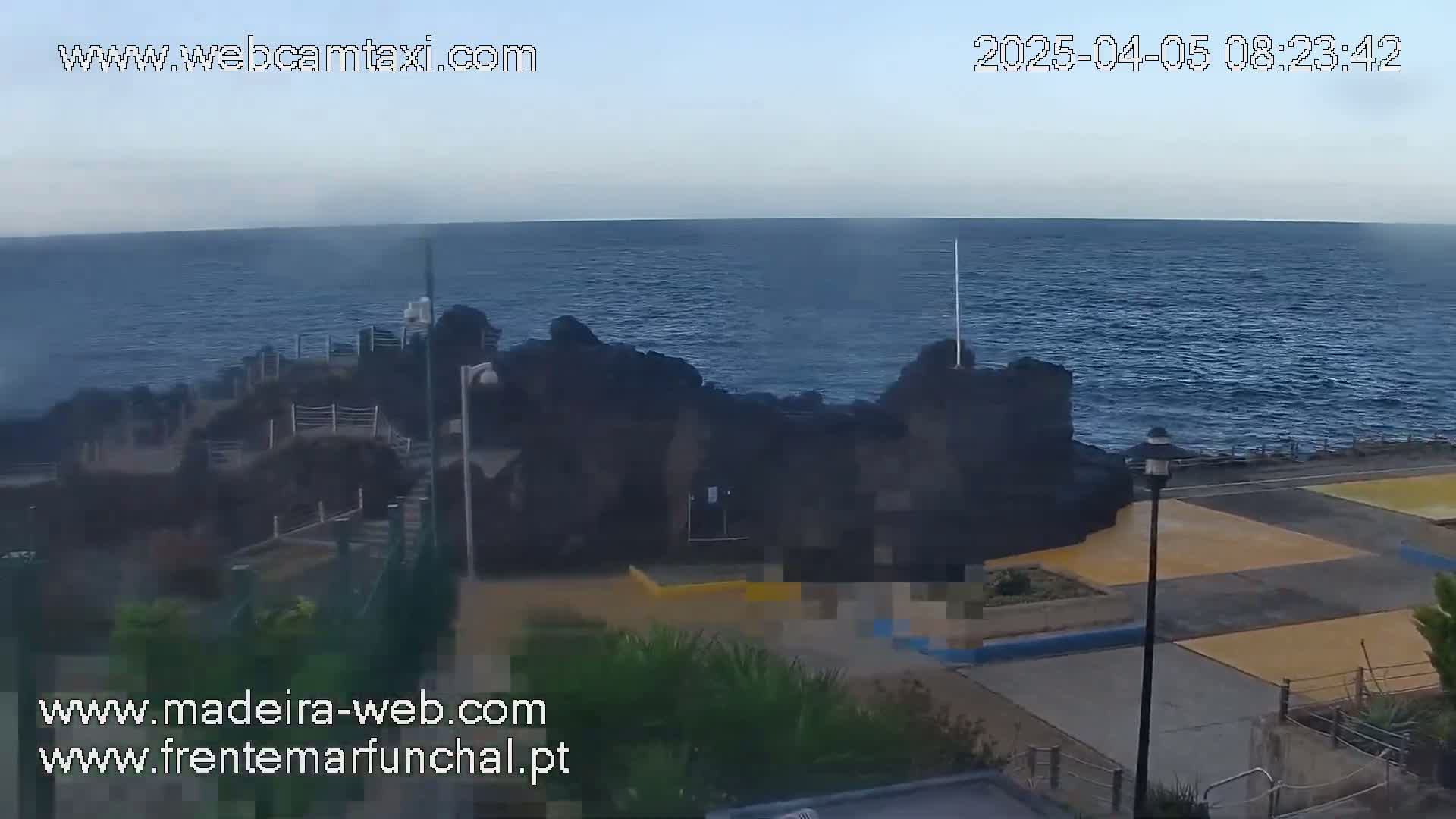 A rocky coastline with calm, dark blue ocean under a clear sky is viewed from above, showing a paved area with yellow sections near the water.