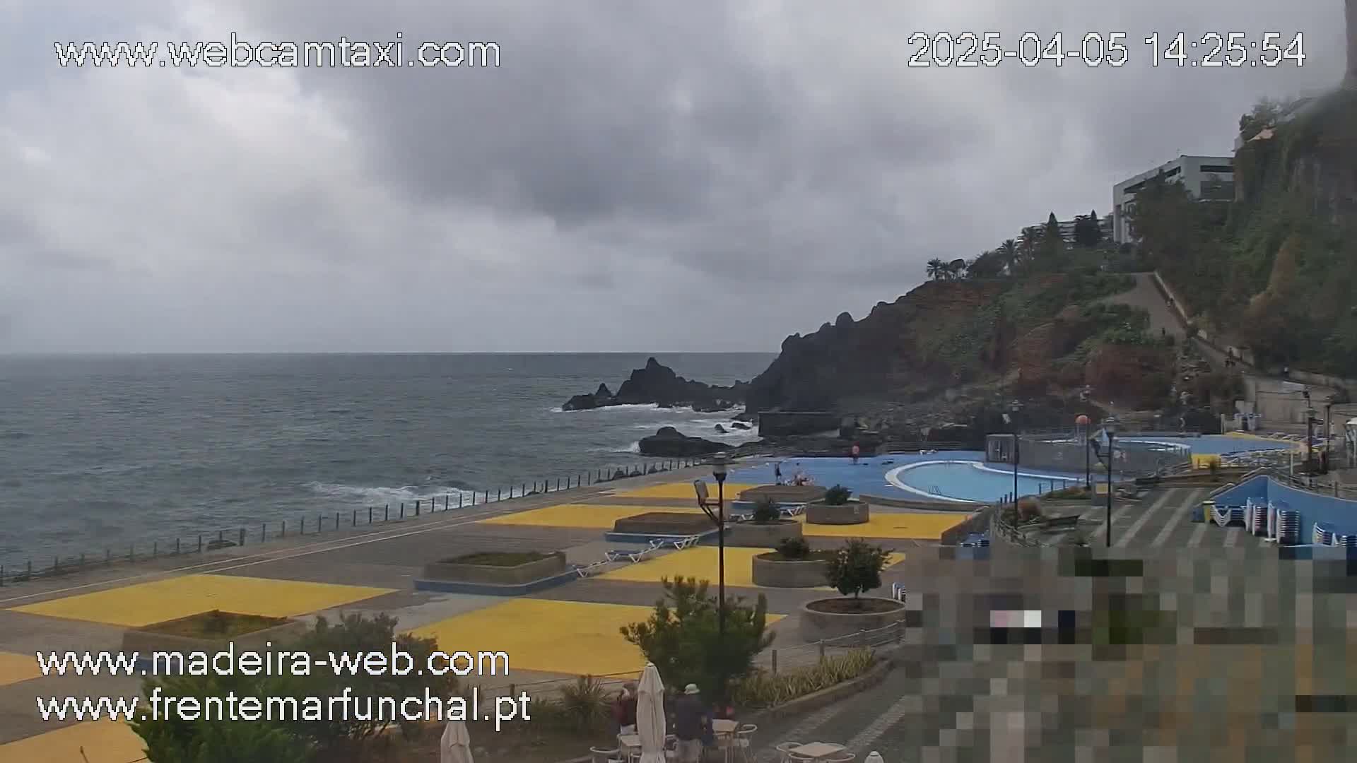 A coastal outdoor scene on an overcast day shows a paved area with yellow sections next to a dark rocky shoreline and ocean, featuring a swimming pool and some people.
