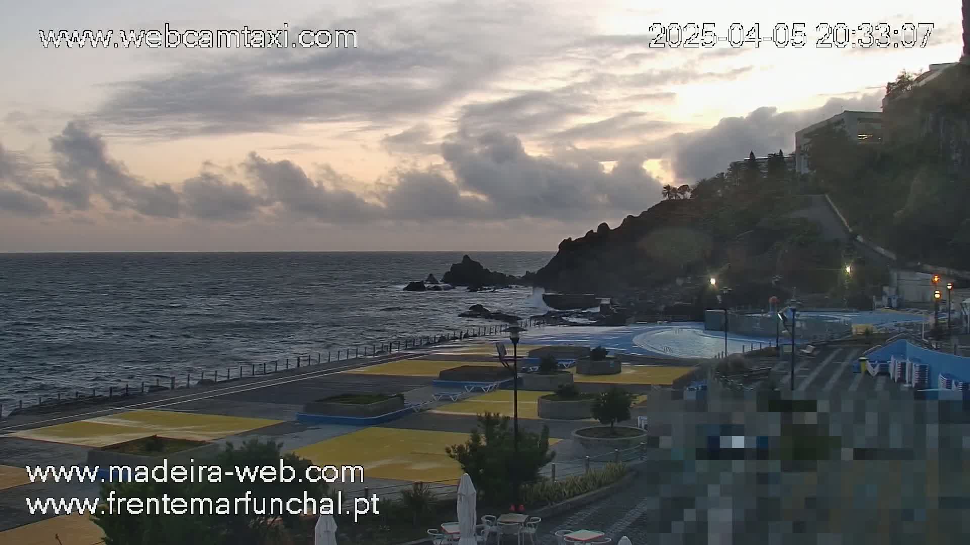 A seaside outdoor scene at dusk shows a partially cloudy sky over dark ocean water, rocky coastline with a building in the background, and a paved area with what appears to be an outdoor swimming pool and seating.