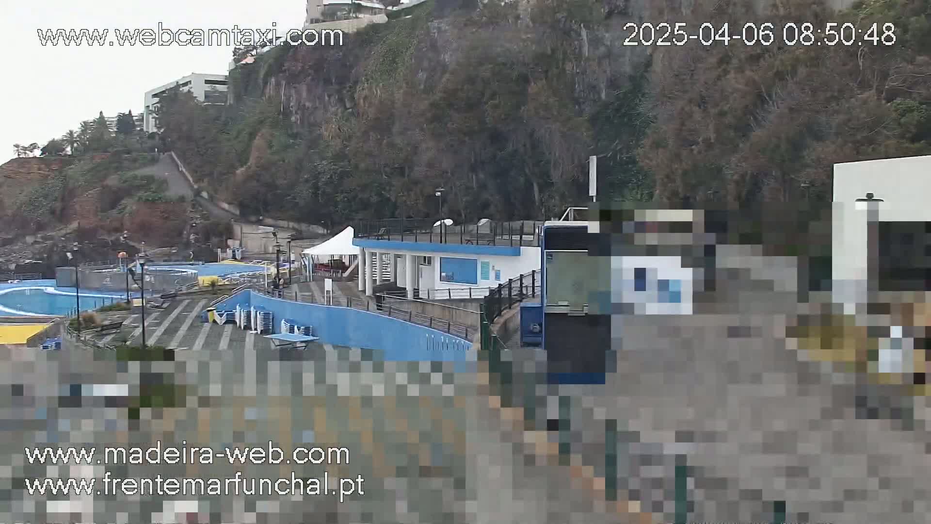 An overcast day reveals an outdoor pool complex situated on a cliffside, with a small building and a cable car partially visible.
