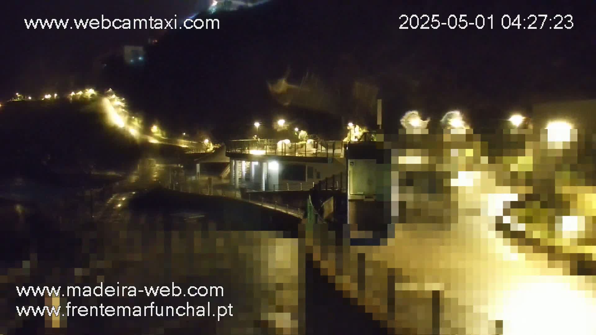 A nighttime view of a rain-slicked waterfront area with illuminated buildings and a partially obscured vessel.