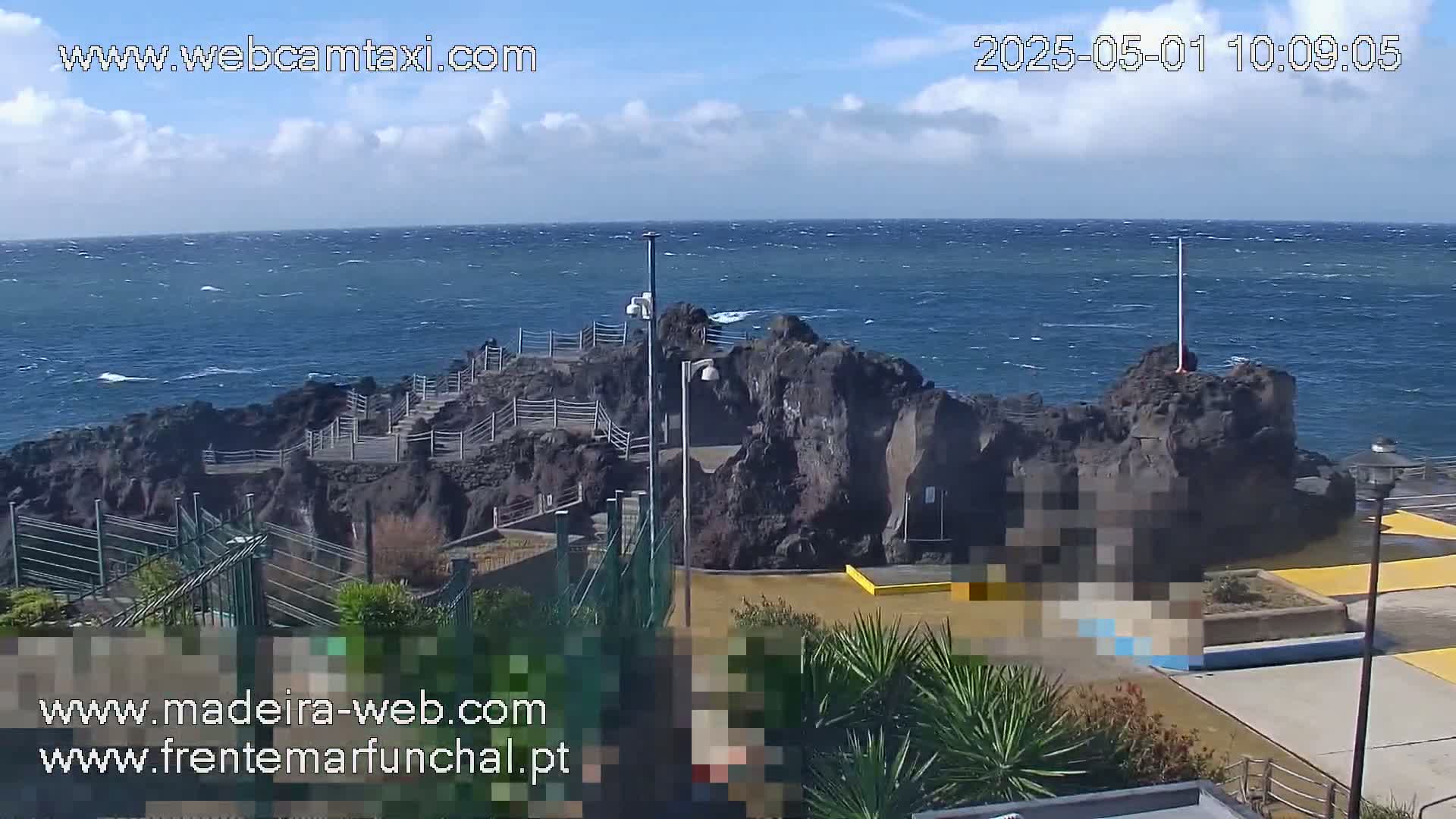 A rocky outcrop with a walkway extends into a wavy, blue ocean under a partly cloudy sky.