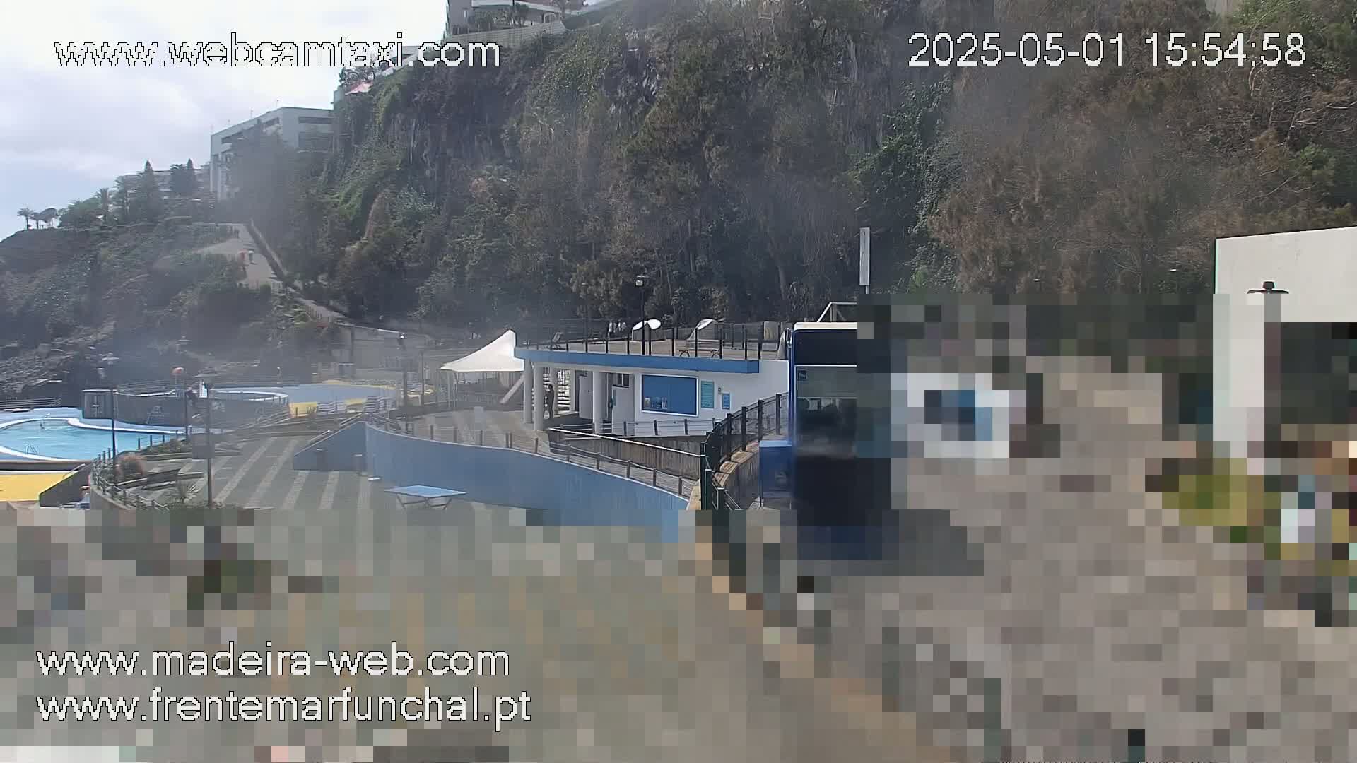 An outdoor scene on an overcast day shows a swimming pool complex nestled into a cliffside, with a bus partially obscuring the view.