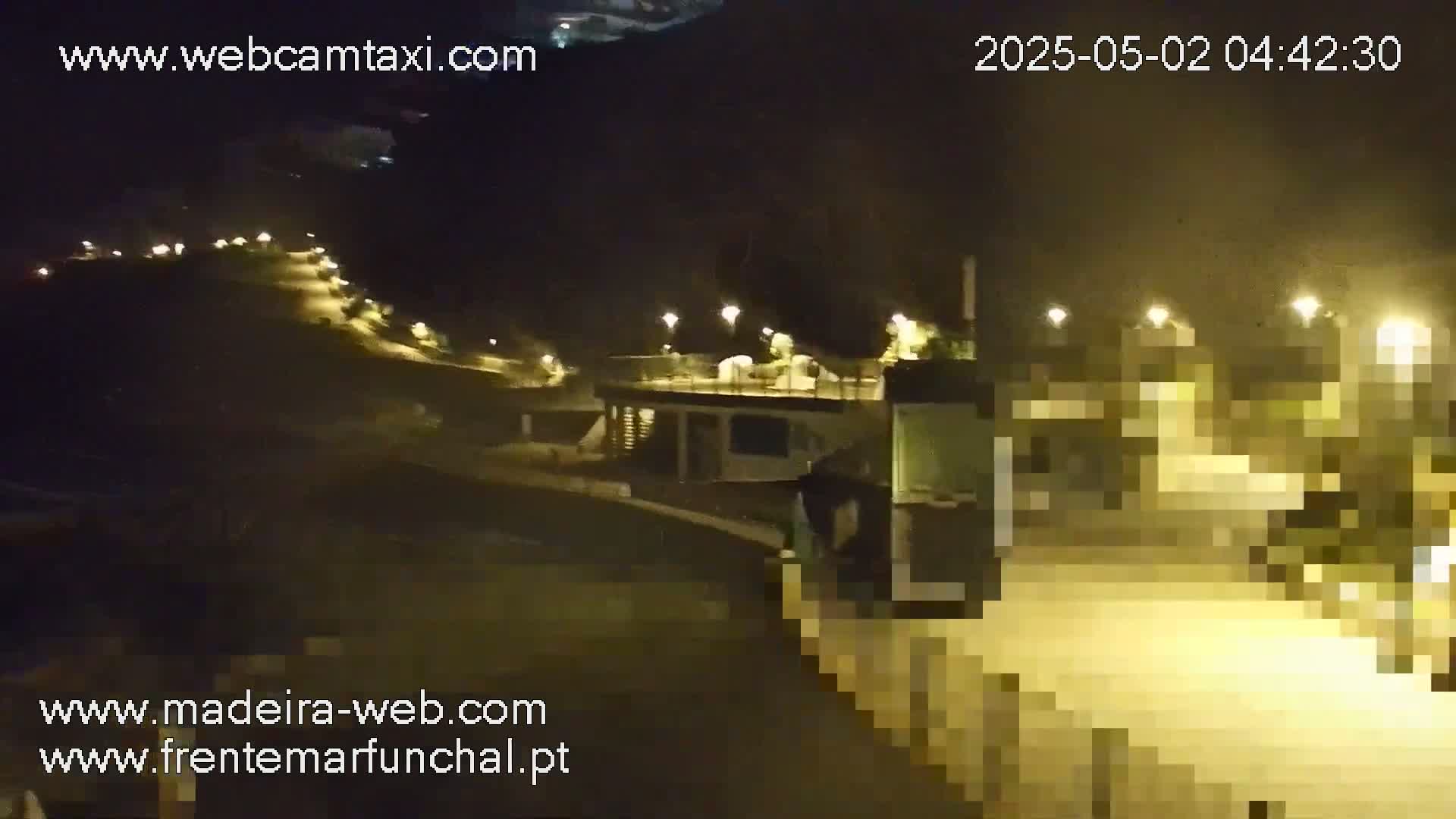 A nighttime, illuminated view of a paved area with buildings and a hillside, possibly a coastal area.