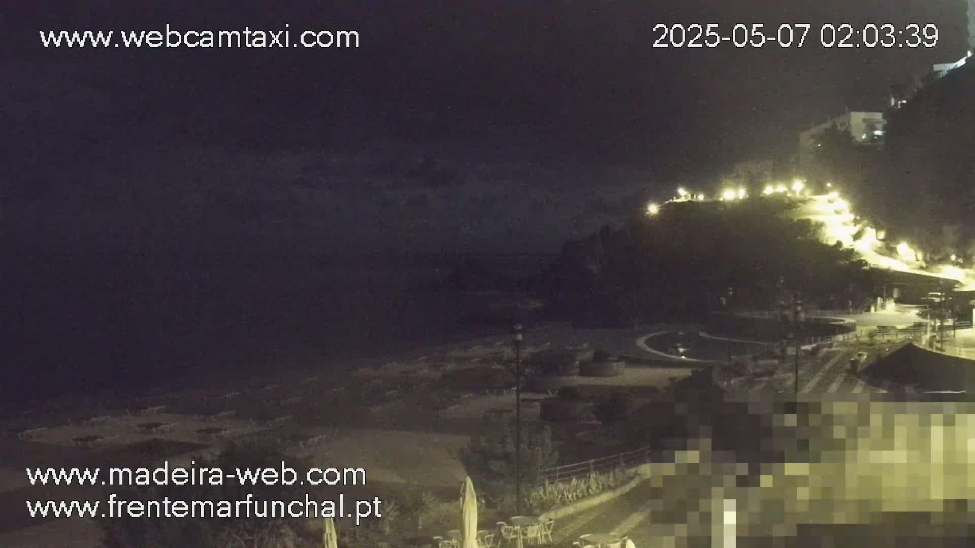 A nighttime beach scene shows a dark ocean, a mostly empty beach with some lounge chairs, and a hill in the background with buildings and lights.