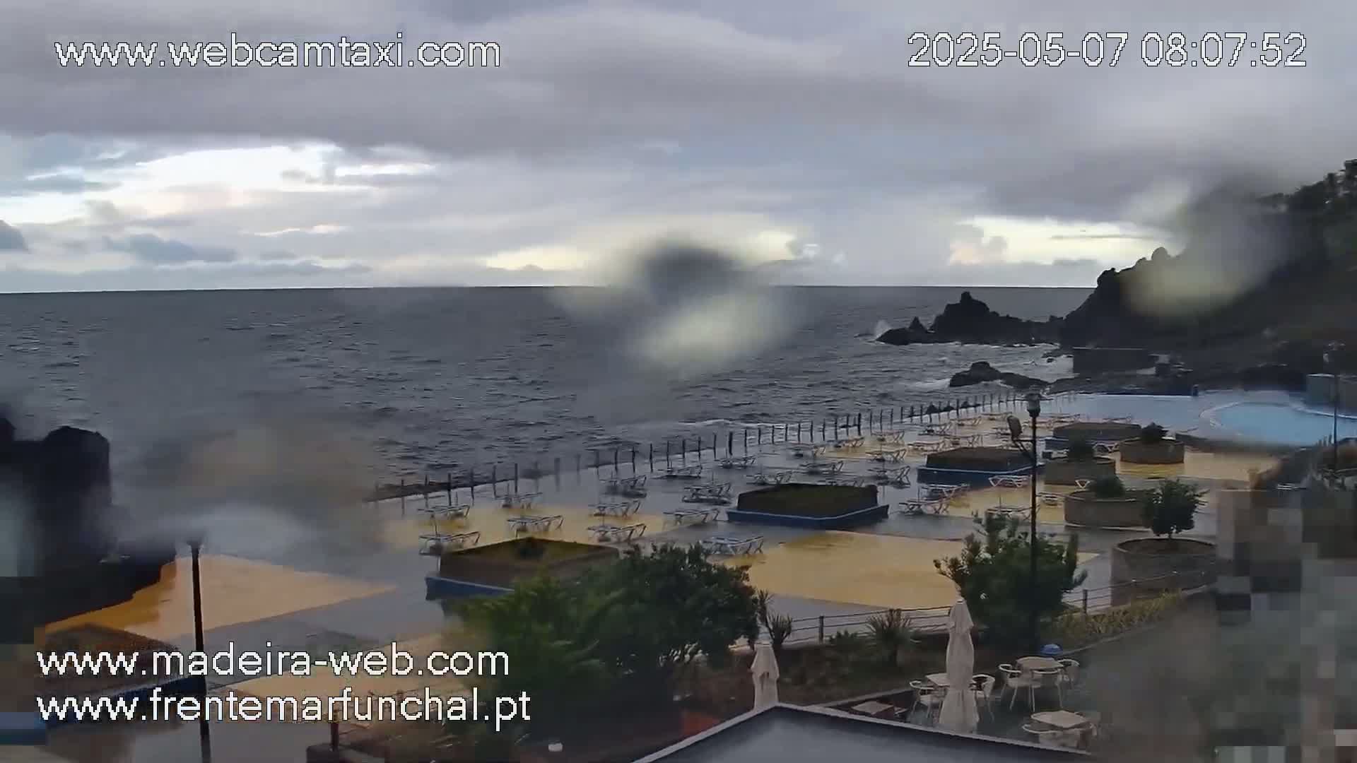 A mostly cloudy day shows a rocky coast with a partially flooded outdoor seating area near a wavy ocean.