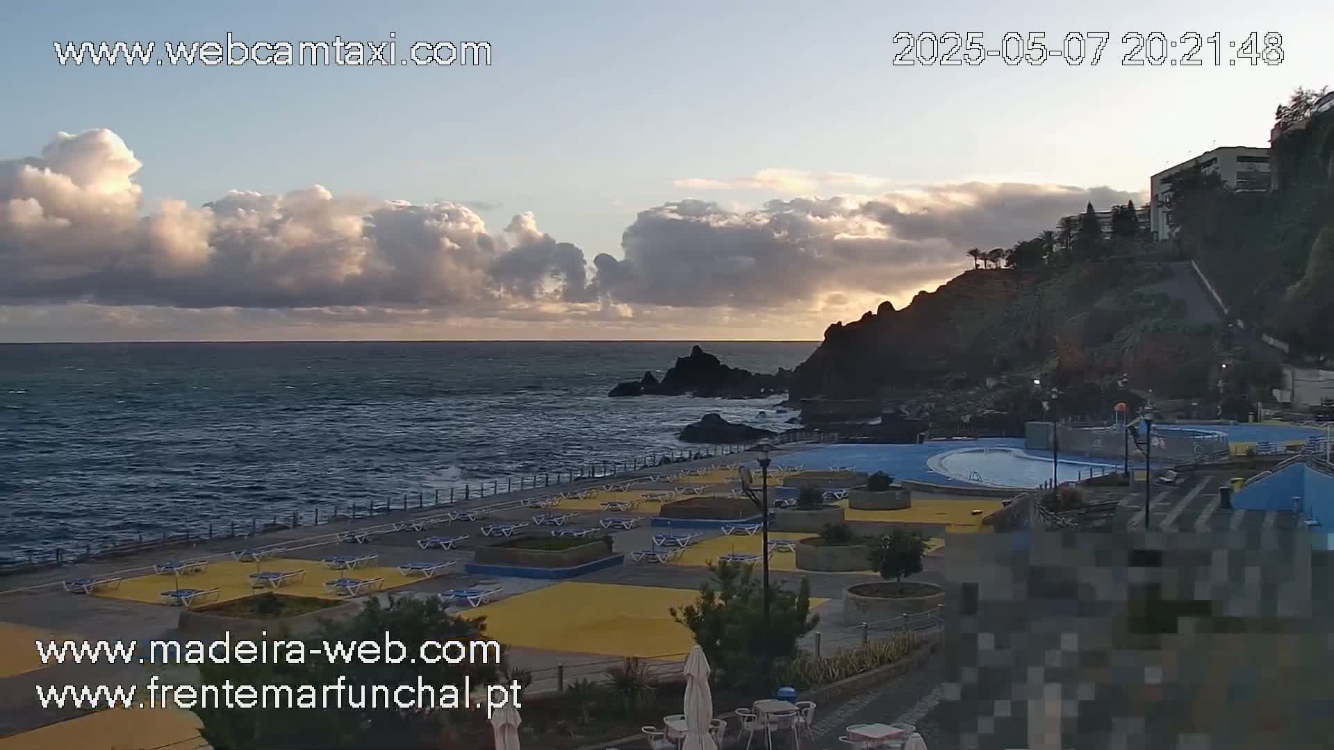 A mostly cloudy sunset over a rocky coastline with a pool and lounge chairs situated on a paved area overlooking the ocean.