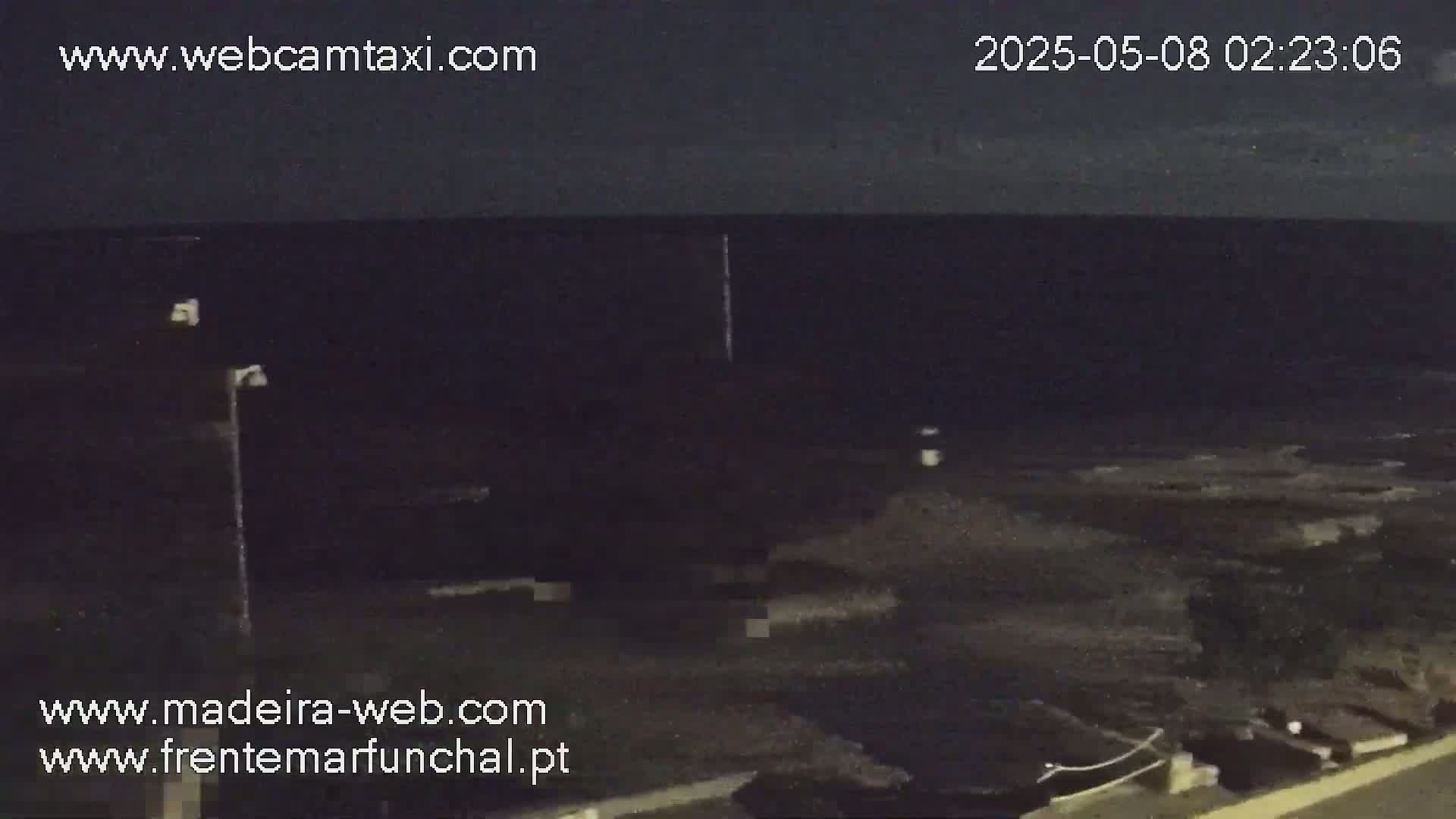 A nighttime, low-light view of a dark ocean with a rocky shoreline, a single lamppost, and some sparse vegetation visible near the water.