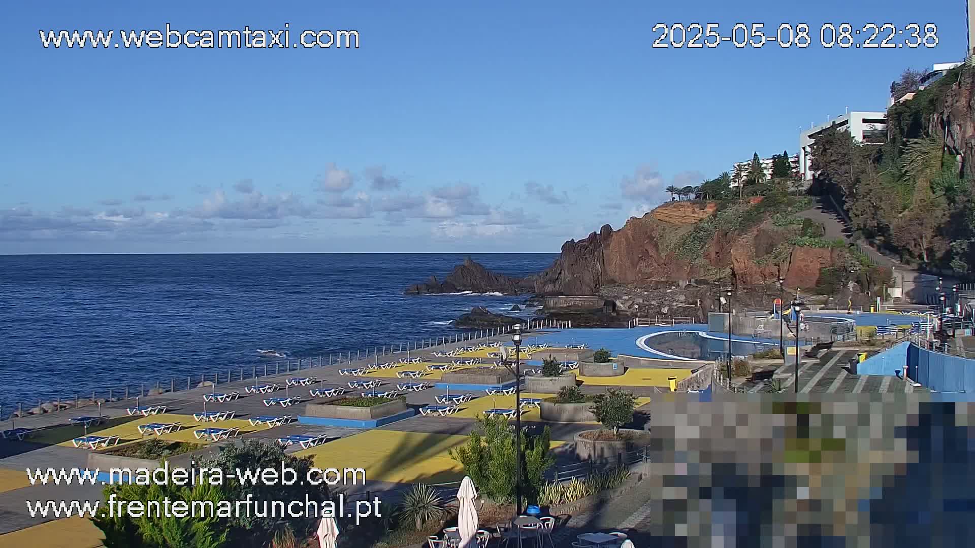 A coastal outdoor scene on a mostly clear day shows a pool and lounge area next to a rocky cliff overlooking a calm ocean.
