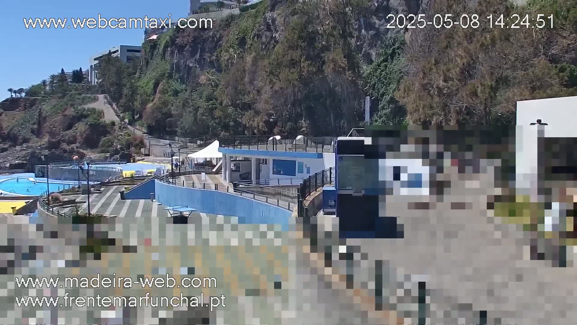 A sunny outdoor scene shows a swimming pool area next to a cliffside, with a bus partially visible and buildings in the background.