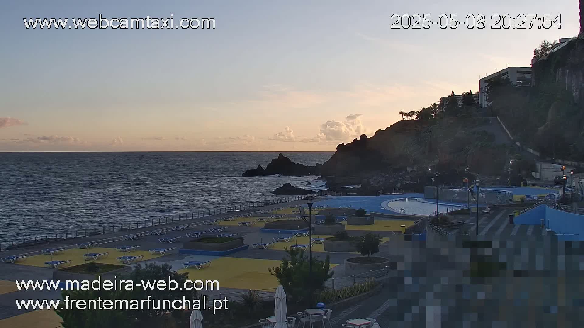 A calm ocean meets a rocky shoreline at sunset, where a paved area with lounge chairs and a pool overlooks the water.