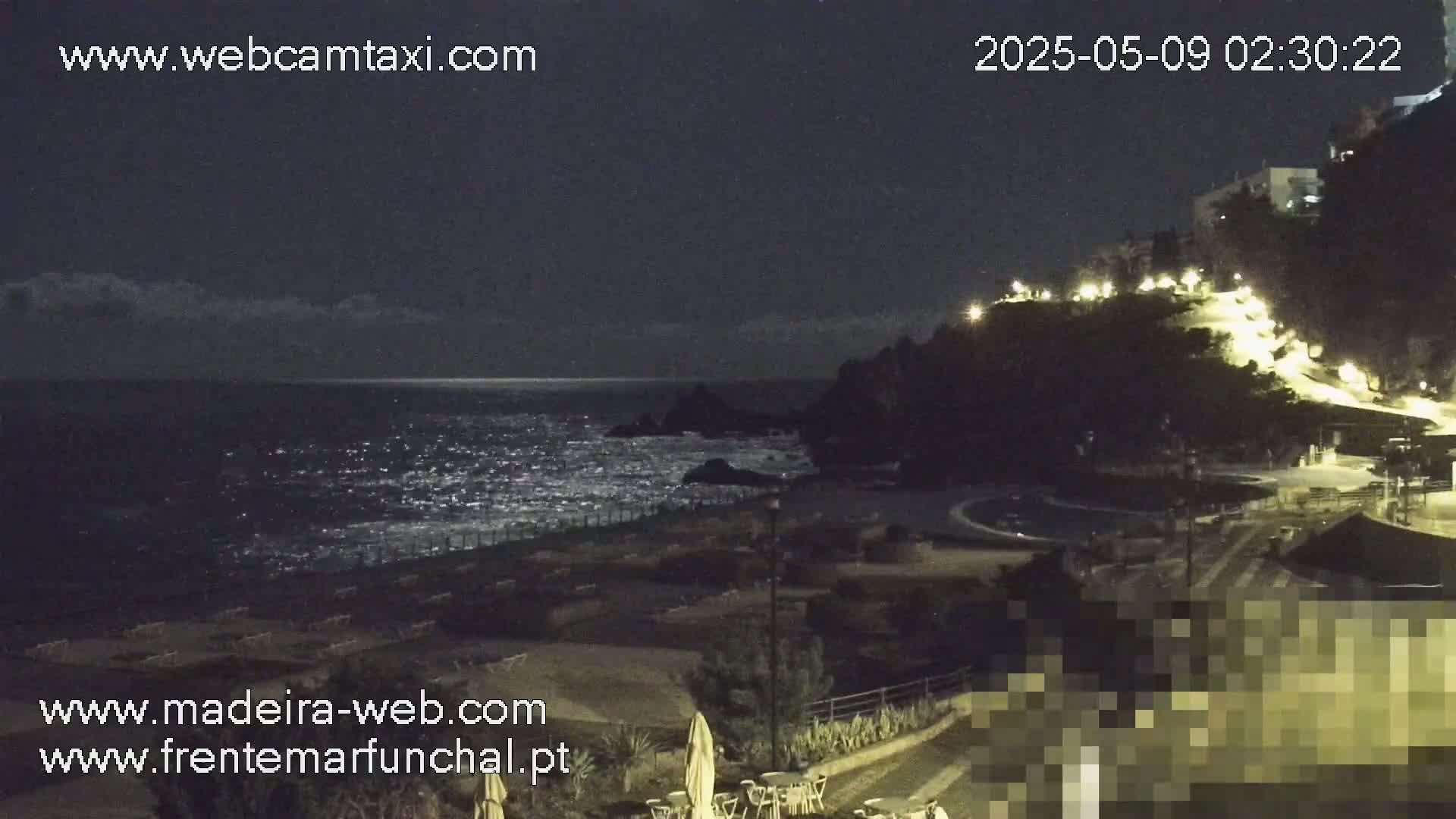 A nighttime coastal scene shows a dark, wavy ocean under a mostly clear sky, a dark beach with empty lounge chairs, and a hilltop illuminated by lights.