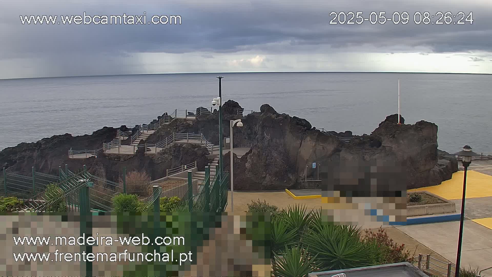 A rocky outcrop with stairs leading to it juts into a calm ocean under a cloudy sky.