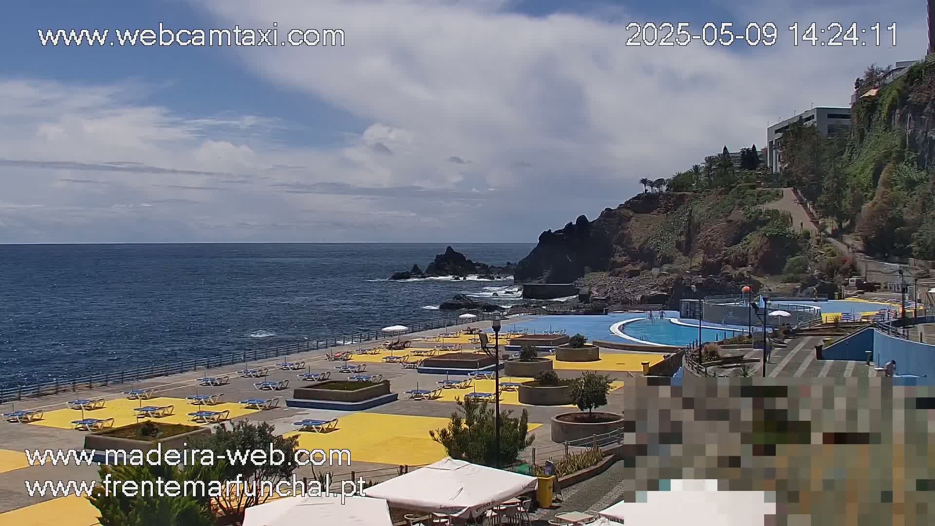 An outdoor scene shows a partly cloudy sky over a blue ocean with a rocky shoreline featuring a swimming pool and sunbathing area with yellow-colored deck chairs.