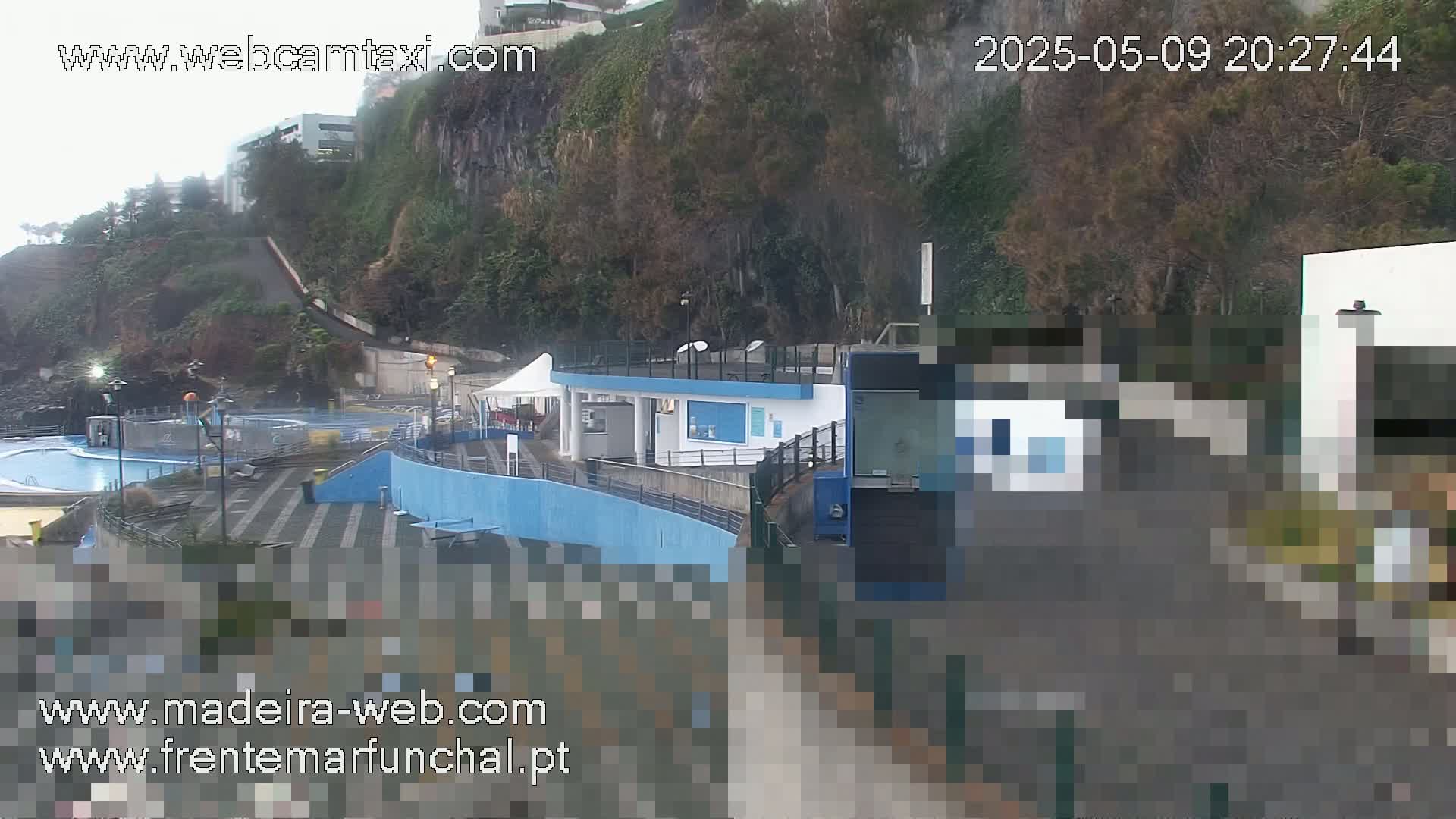 An outdoor scene on an overcast day shows a building complex near a cliffside with a small swimming pool, and a paved area.