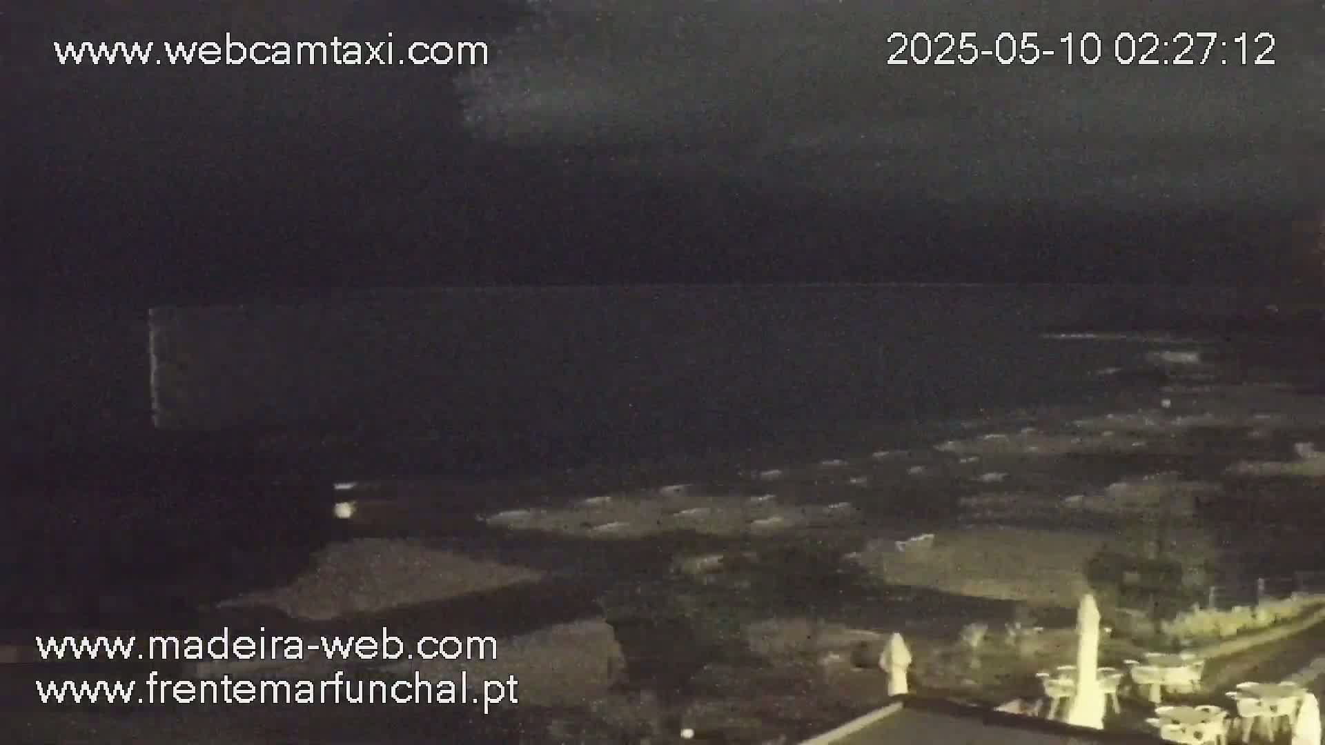 A nighttime view of a dark ocean and a shoreline with outdoor furniture under a cloudy sky.