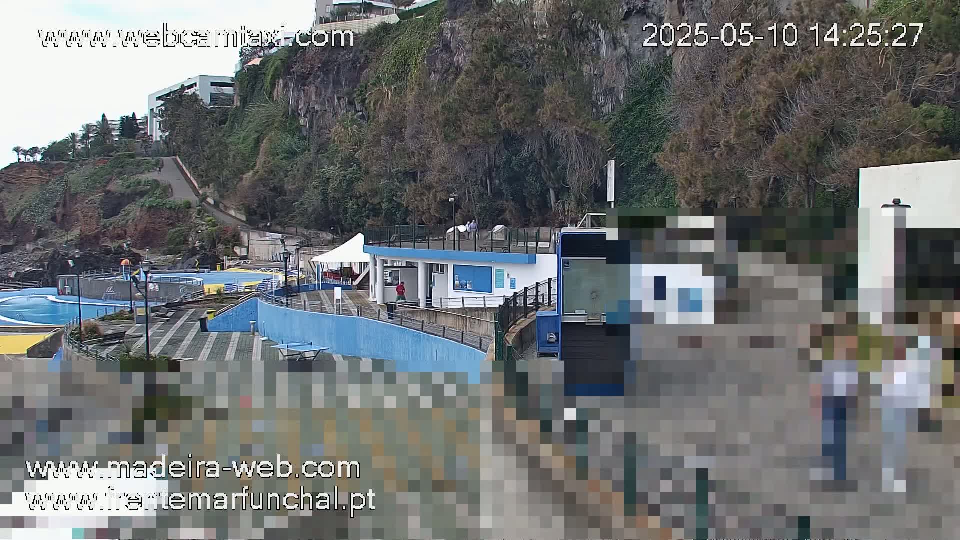 An outdoor scene shows a seaside pool area with a building and a walkway, all situated on a cliffside with trees and overcast skies.