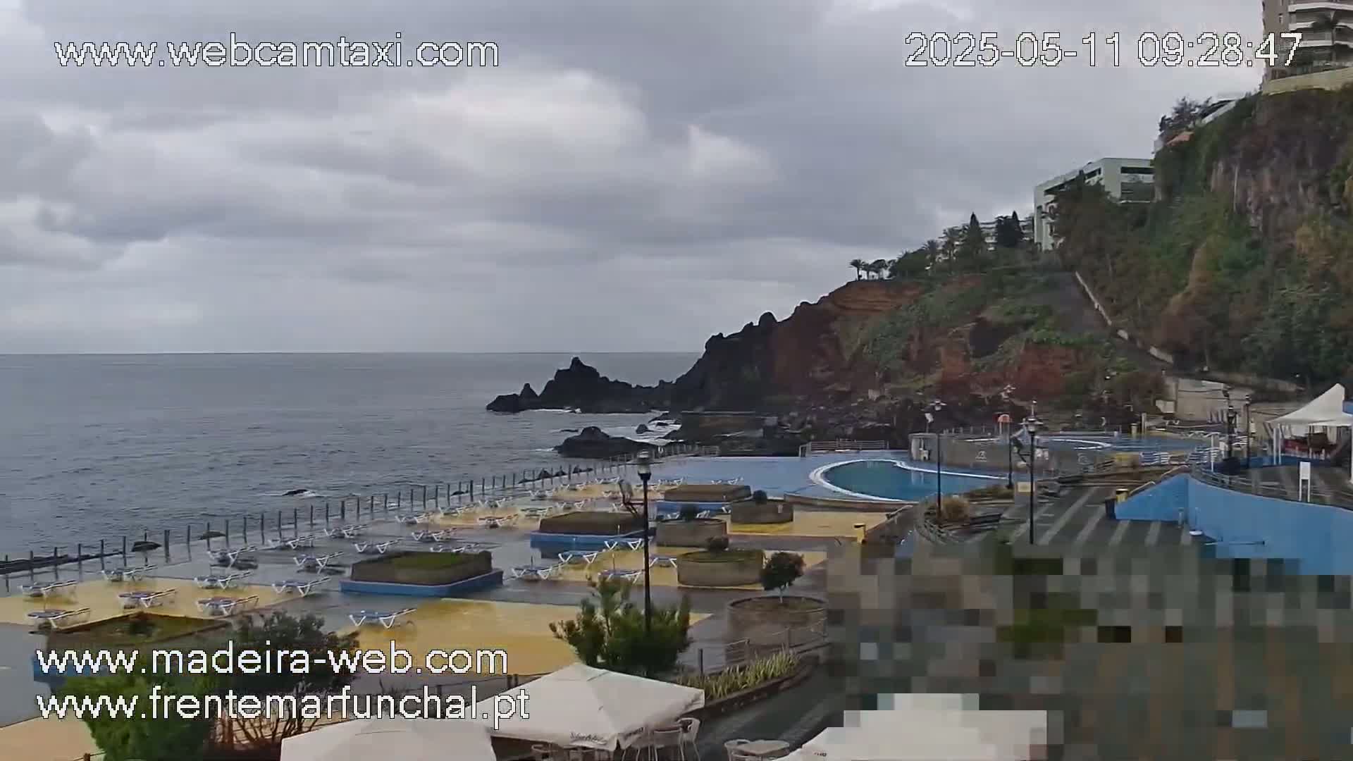 An oceanside pool area with overcast skies sits below a cliff with buildings on top.