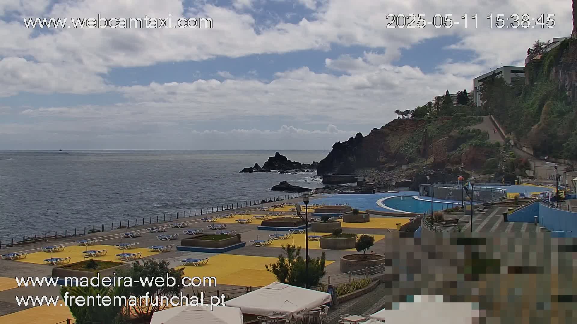 A partly cloudy day shows an outdoor swimming pool area next to the ocean, with lounge chairs and umbrellas set up on a yellow deck, and a rocky cliffside visible in the background.