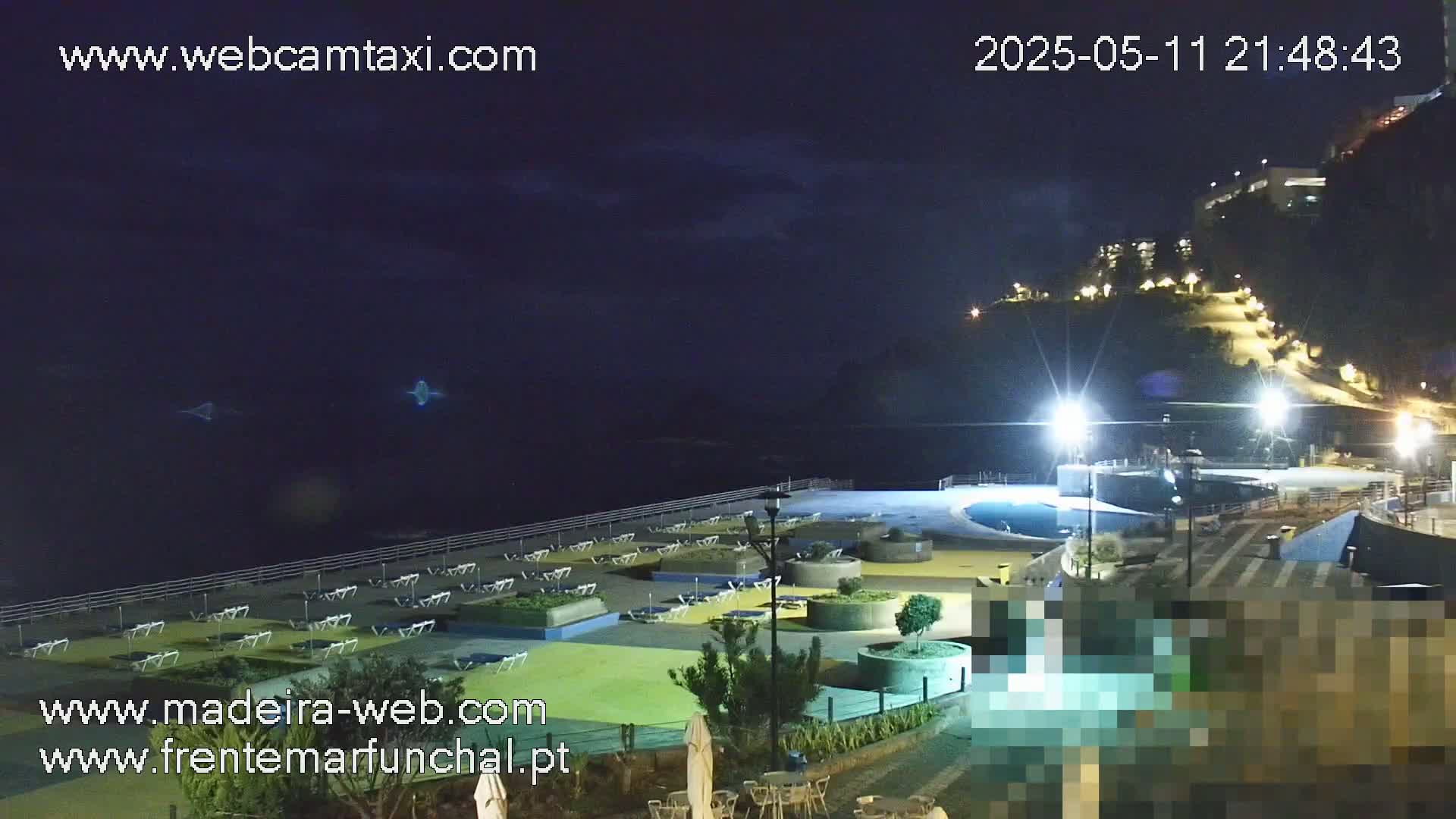 A nighttime view of a coastal area shows a pool and lounge chairs near the ocean under a mostly clear, dark sky.
