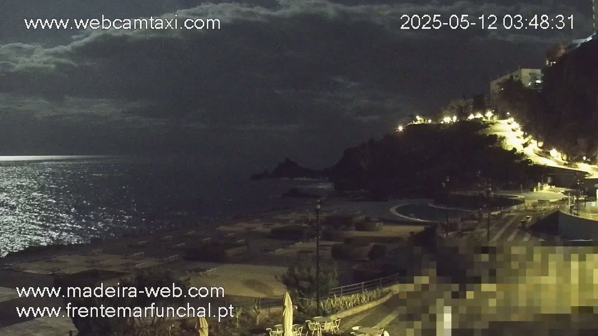A nighttime coastal scene shows a dark ocean under a partly cloudy sky, with a dimly lit beach and a hilltop with illuminated pathways in the background.