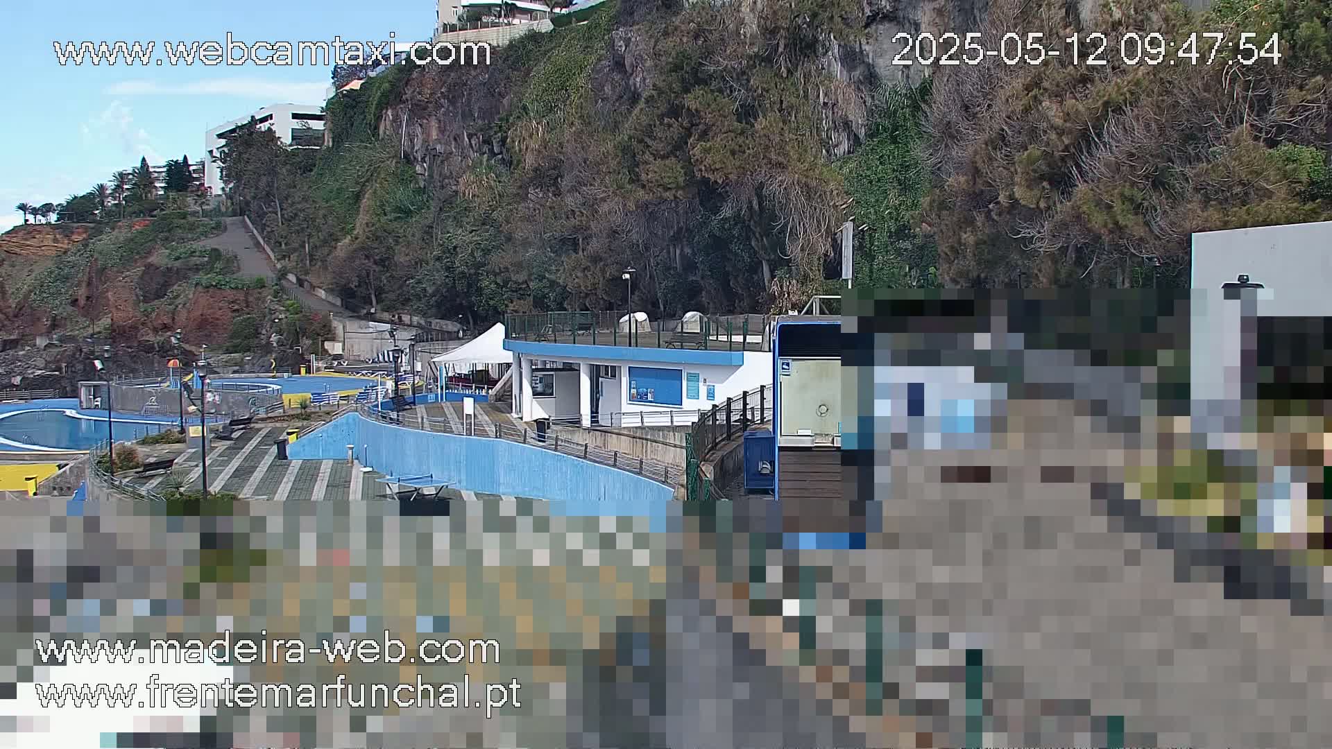 An outdoor scene shows a pool complex situated on a cliffside overlooking the ocean under a mostly clear sky.