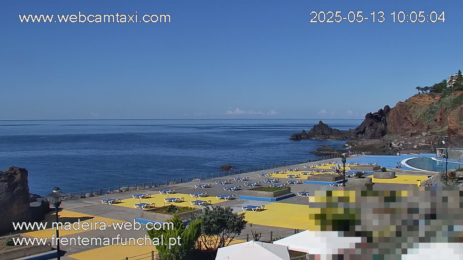 An outdoor view shows a mostly sunny day overlooking a calm ocean with a seaside area featuring lounge chairs and a swimming pool next to a rocky cliff.