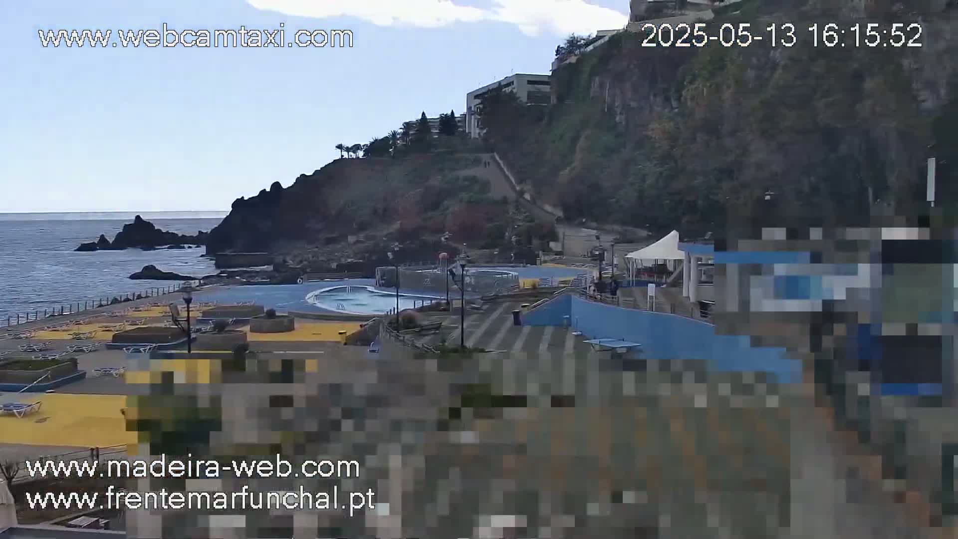 An outdoor view of a seaside swimming pool area with a partly cloudy sky, situated below a cliff with buildings.