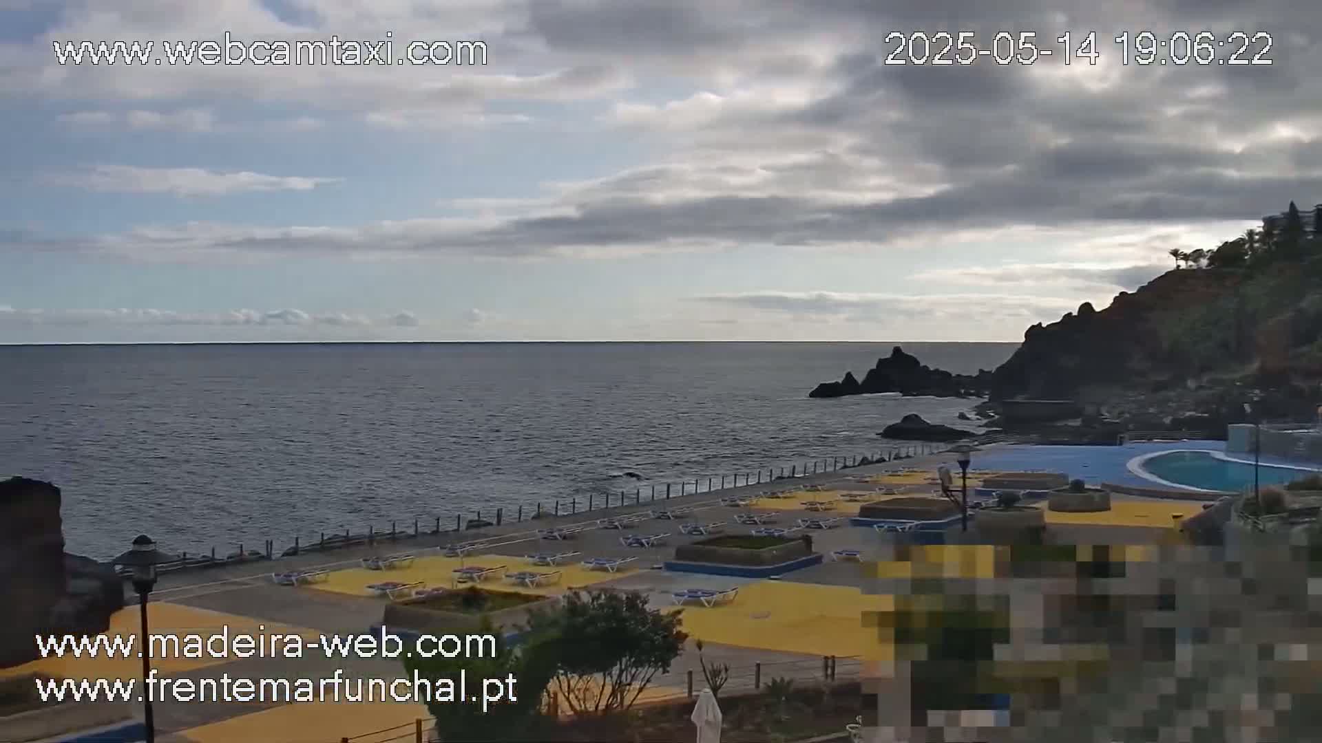 An outdoor scene shows a partially cloudy sky over a dark-blue ocean, a rocky shoreline with a pool and yellow lounge chairs.