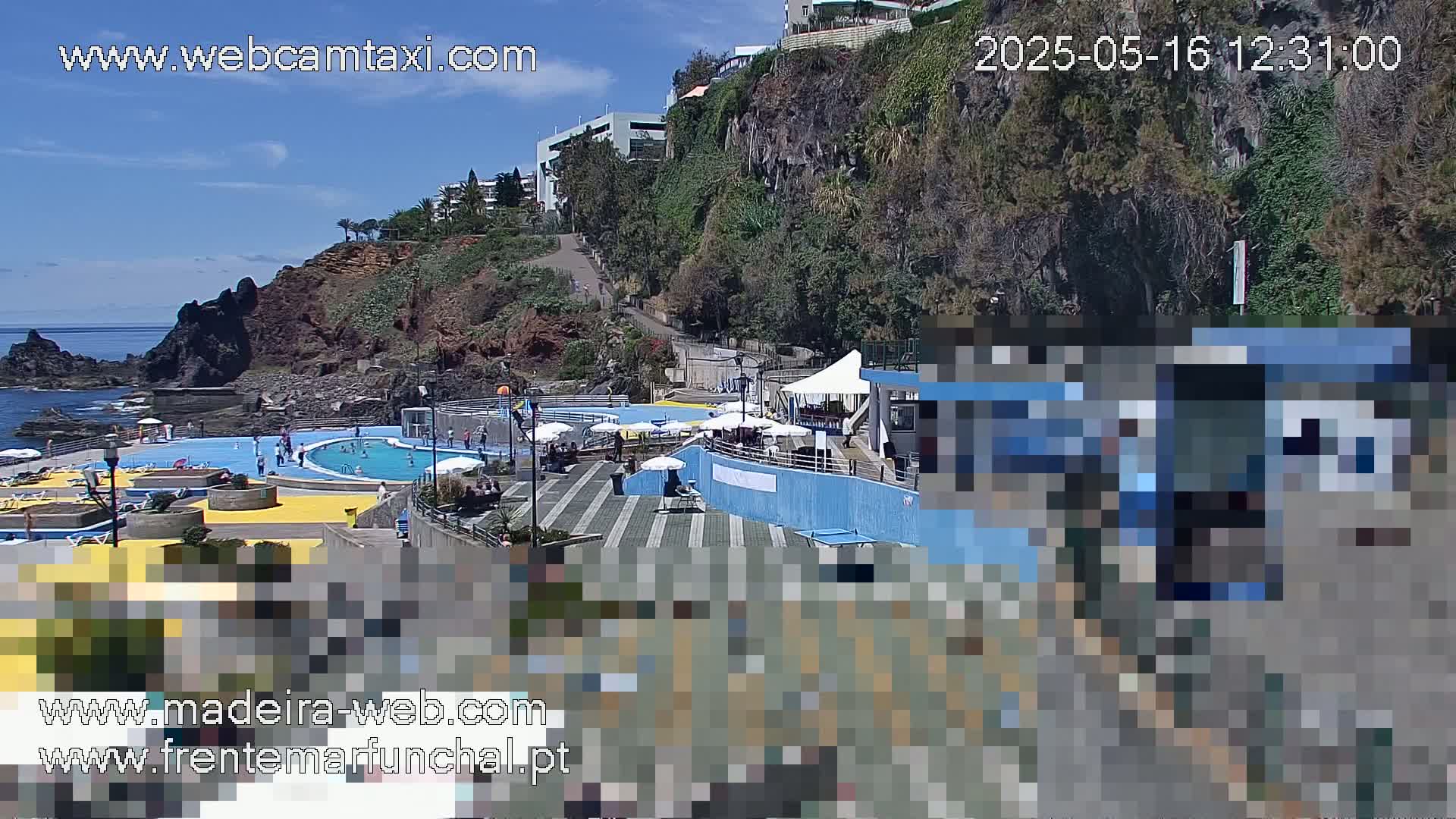 An outdoor scene on a sunny day shows a seaside pool area with people swimming and relaxing, situated on a rocky cliffside with buildings and vegetation visible in the background.
