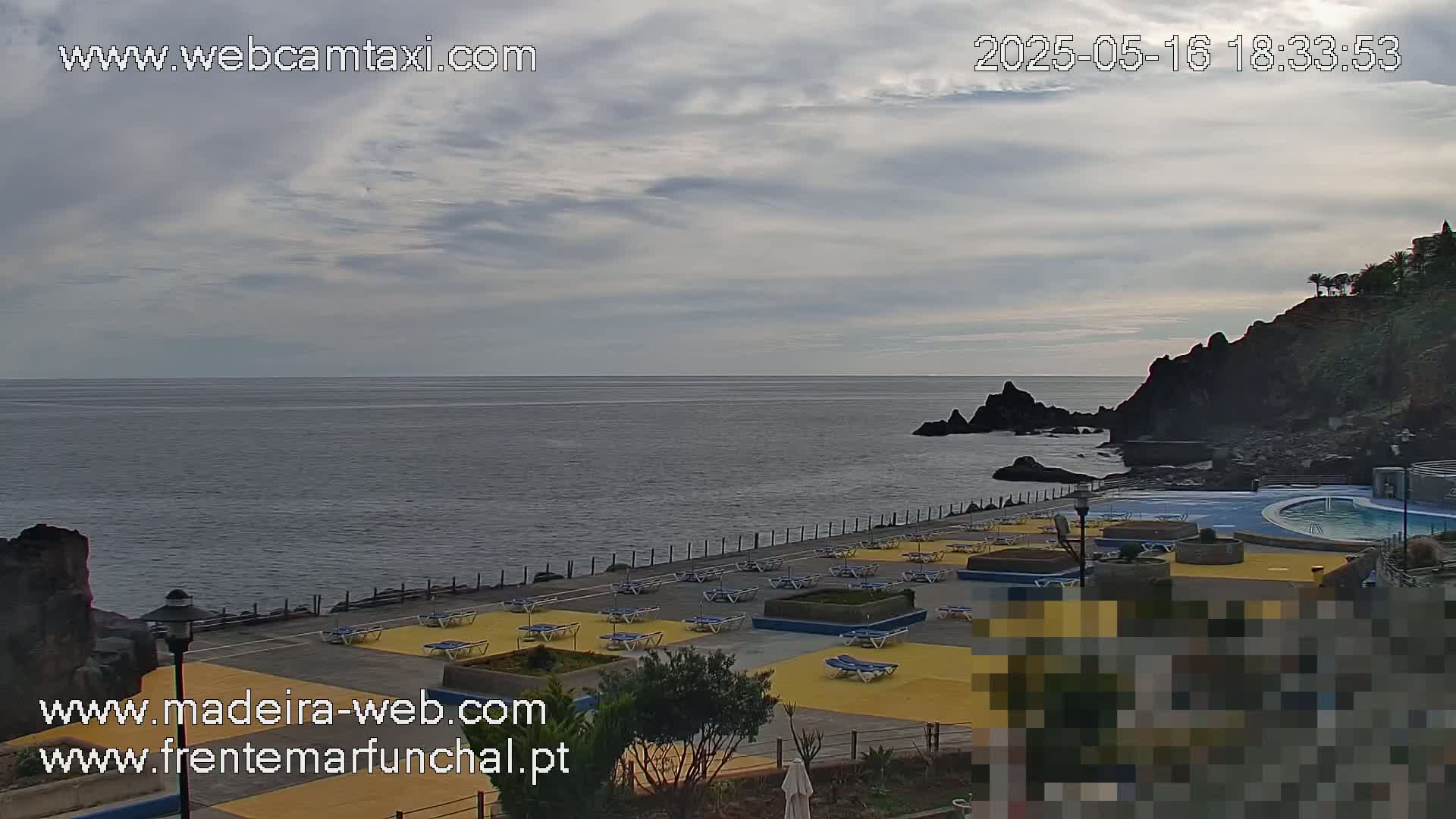 An outdoor scene shows a mostly cloudy sky over a calm ocean, with a rocky shoreline featuring a pool area and numerous lounge chairs on a yellow deck.