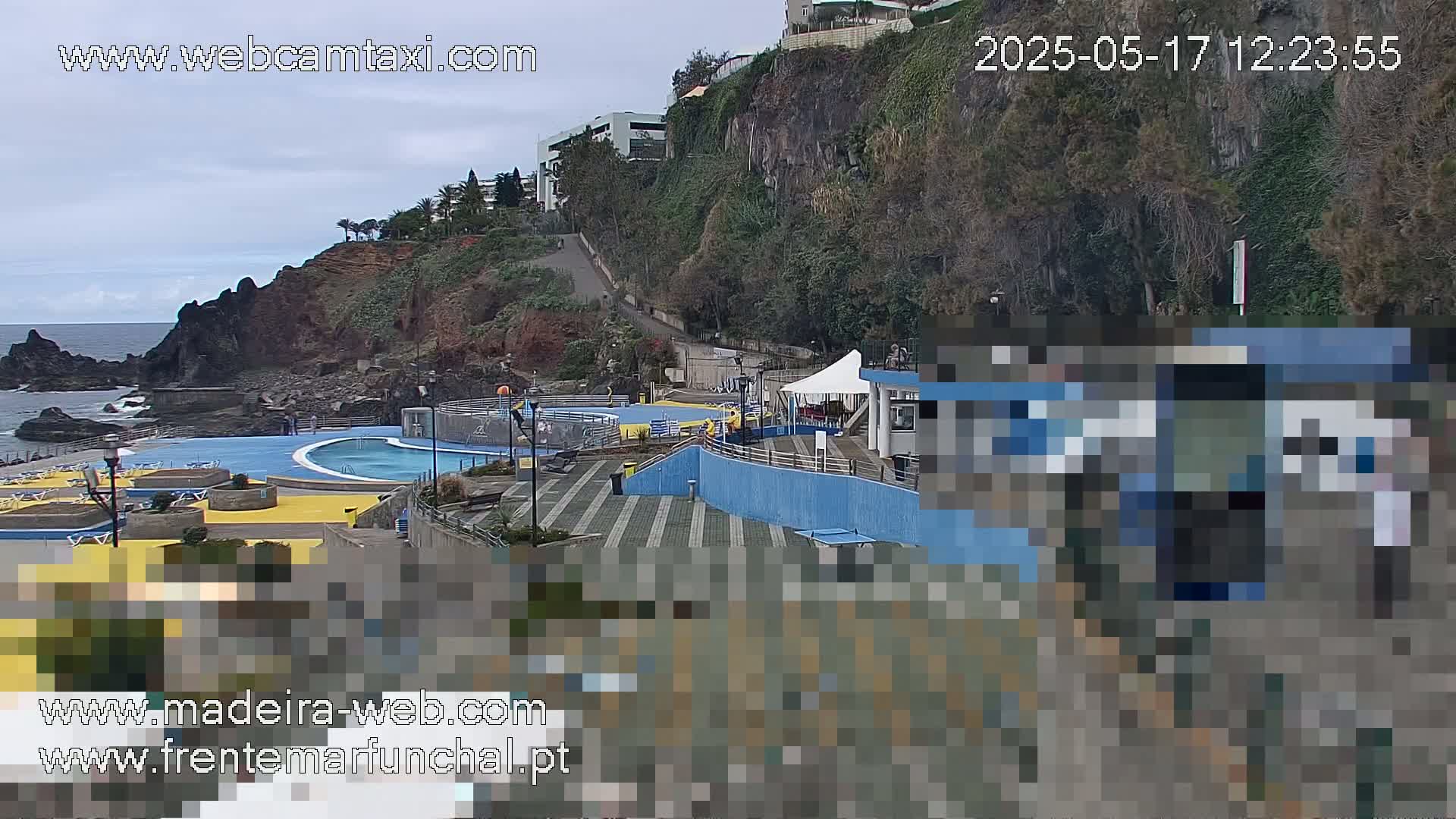 An outdoor pool complex sits on a rocky cliffside overlooking the ocean under an overcast sky.
