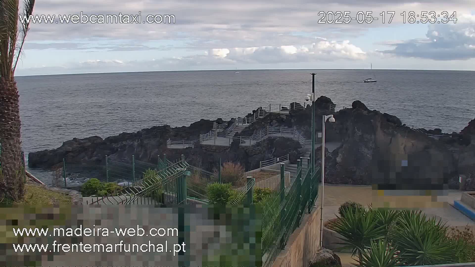 A mostly cloudy day shows a rocky coastline with stairs leading down to the ocean, partially enclosed by a green metal fence, and a small boat visible in the distance.