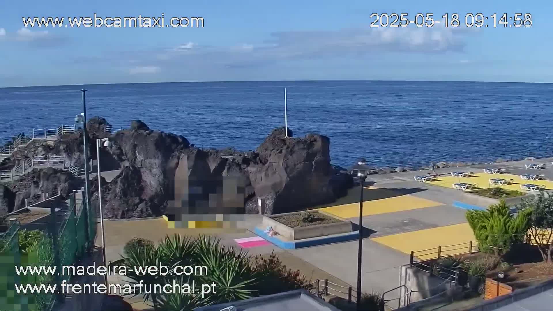 A rocky outcrop juts into a calm, blue ocean under a mostly clear sky, with a paved area containing yellow-painted sections and outdoor furniture.