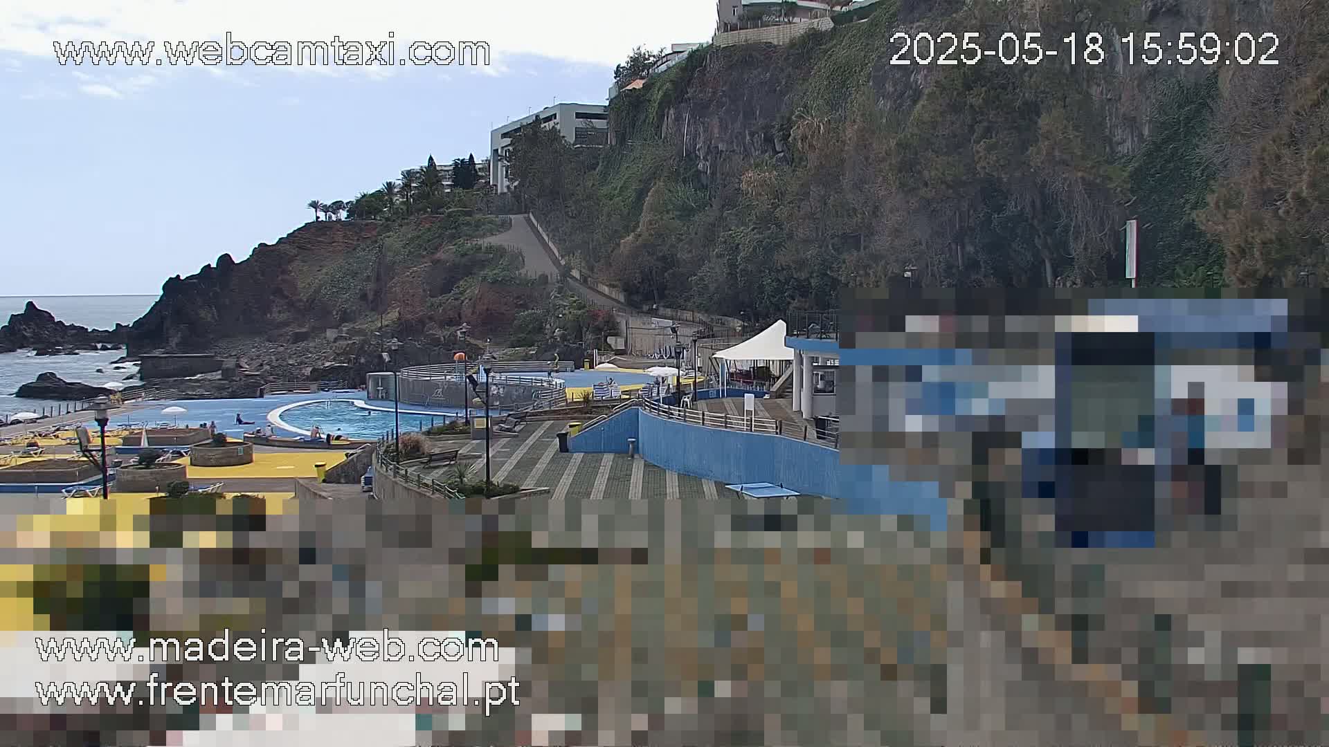 An outdoor scene on a partly cloudy day shows a seaside pool complex with a rocky cliff and buildings in the background.