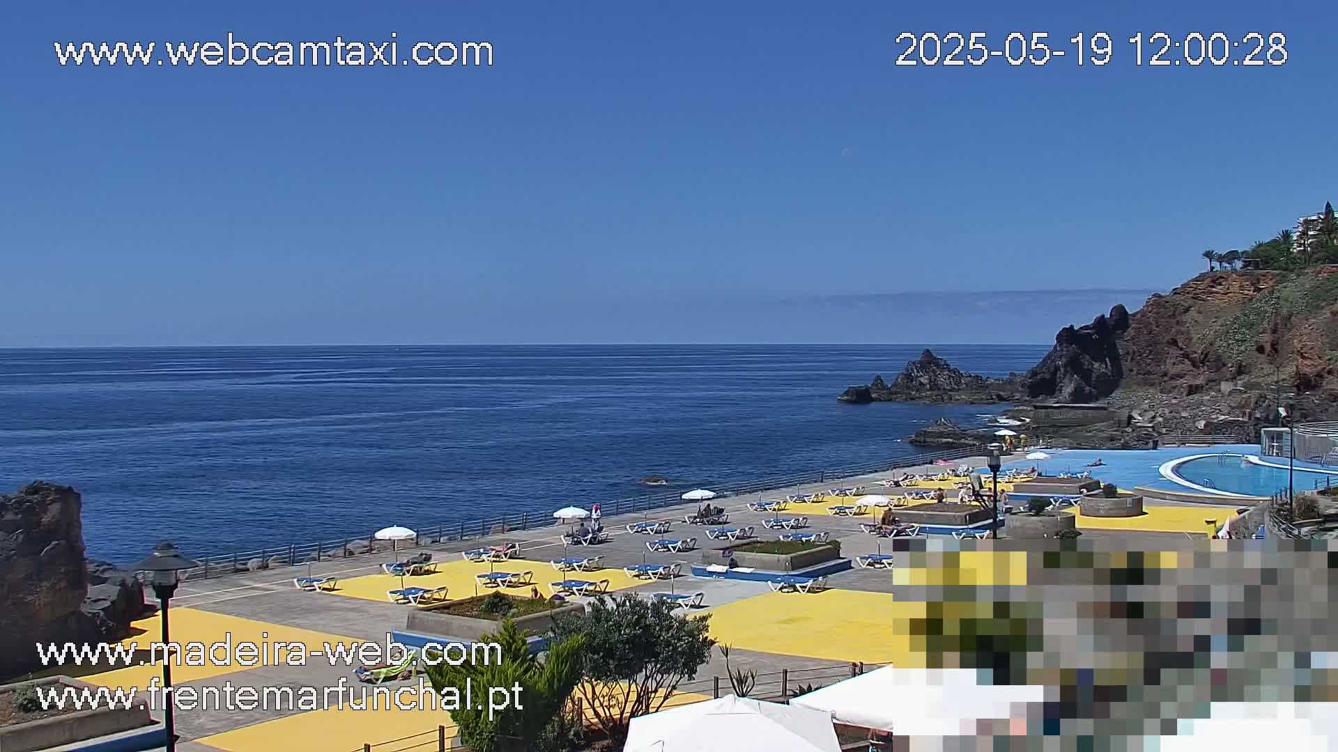An outdoor oceanside scene on a sunny day shows a yellow-tiled area with lounge chairs and umbrellas surrounding a swimming pool, adjacent to a rocky coastline and calm blue ocean.