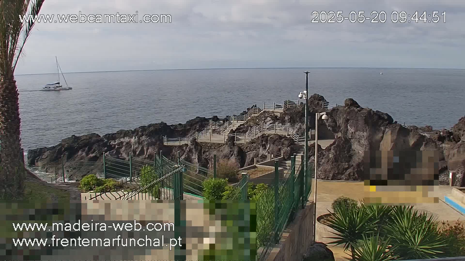 A sailboat is visible on a calm ocean, beyond a rocky coastline with stairs and railings leading down to the water under partly cloudy skies.
