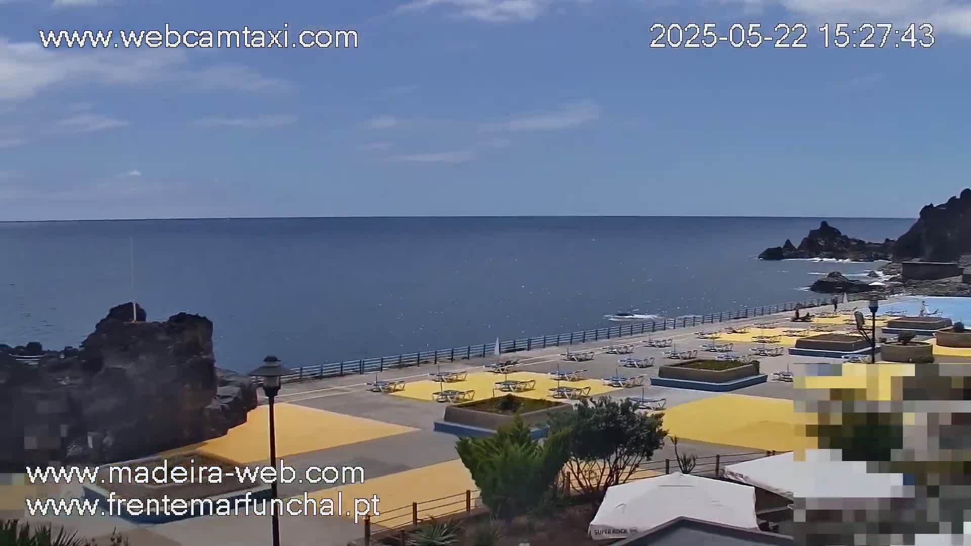 A seaside outdoor scene shows a mostly sunny day with a calm ocean, a paved area with lounge chairs and umbrellas, and dark volcanic rock formations.