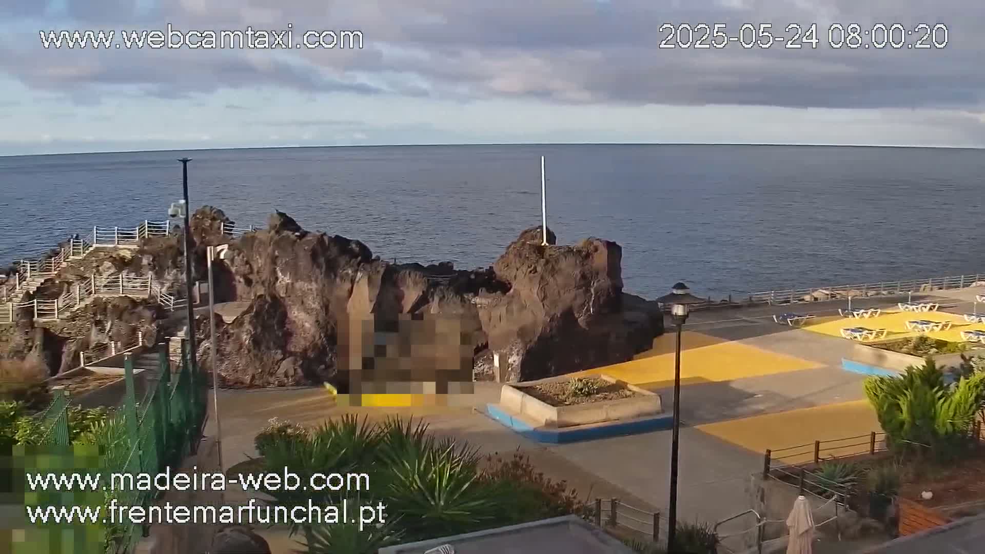 A partly cloudy day reveals a rocky coastal area with a paved walkway, lounge chairs, and ocean views.