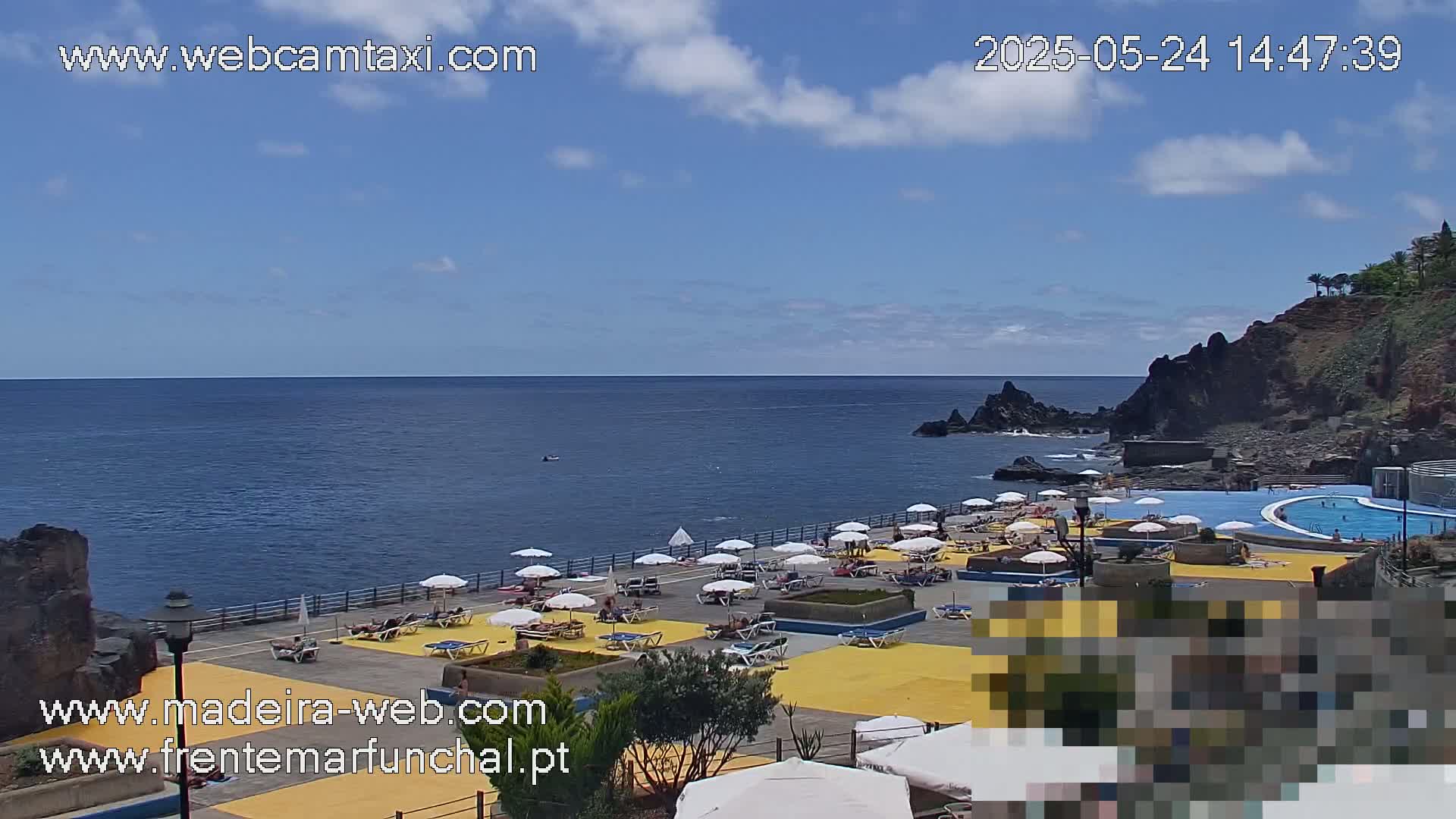 A partly sunny day shows an outdoor swimming pool and sunbathing area next to a rocky ocean coast.