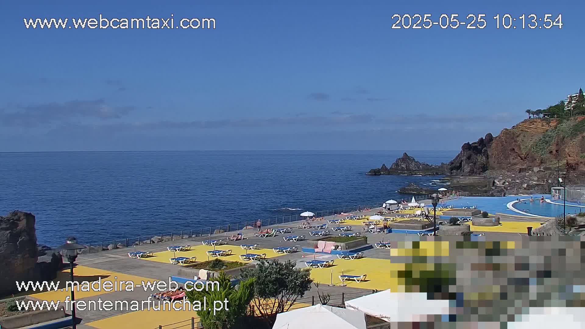 A seaside area with a large pool and numerous lounge chairs on a yellow surface, under a clear blue sky.