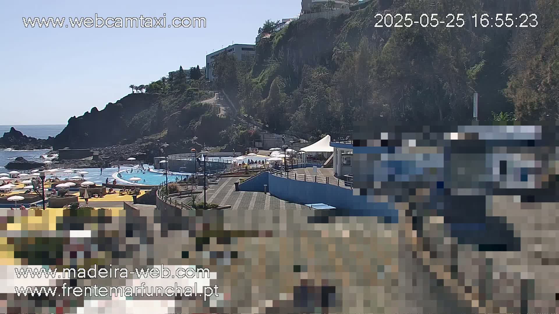 An outdoor scene on a sunny day shows a seaside pool area with people relaxing and swimming, situated on a rocky coast next to a cliffside with buildings.