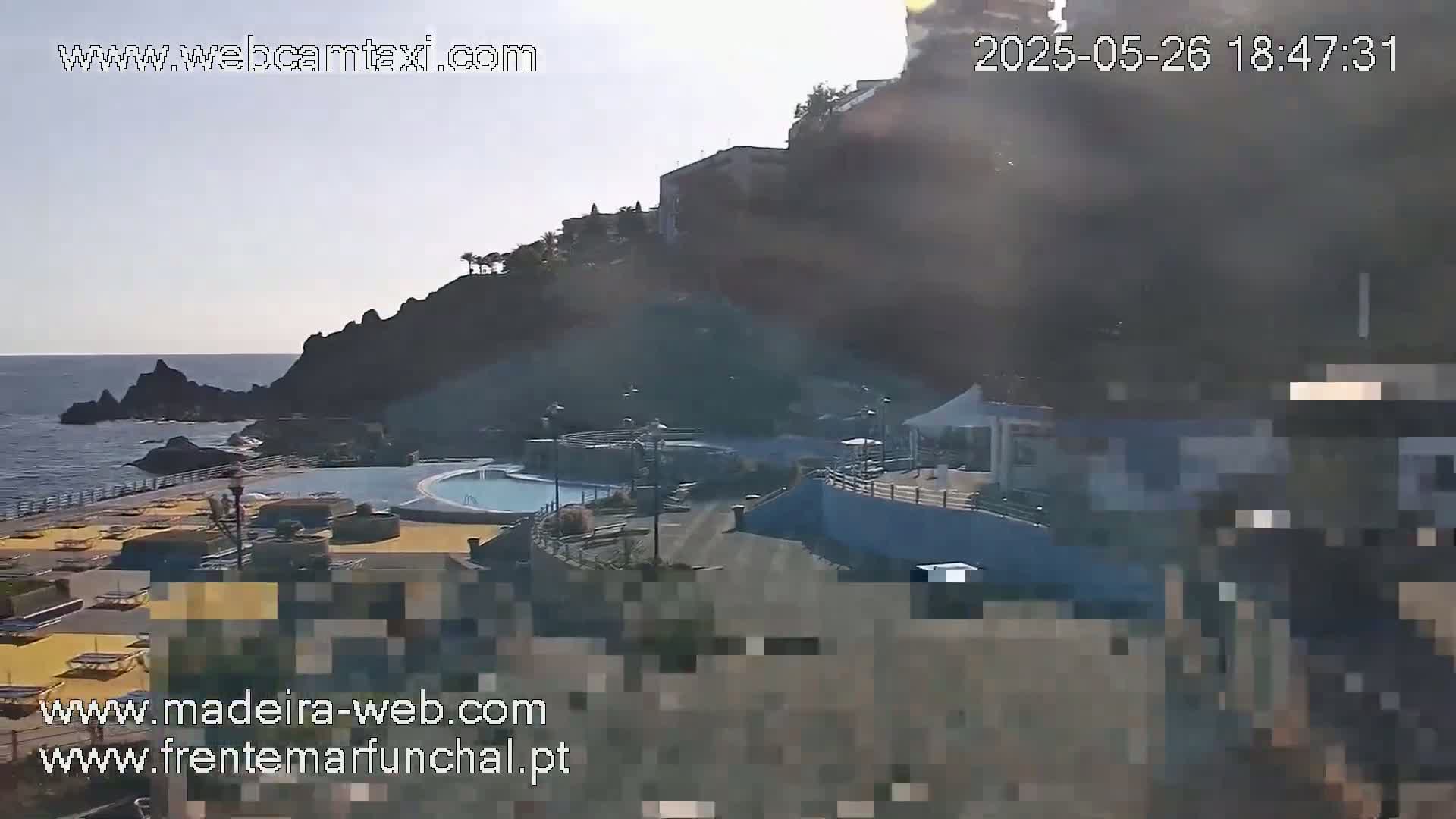 An outdoor view of a seaside pool area and a rocky cliffside on a mostly clear day.