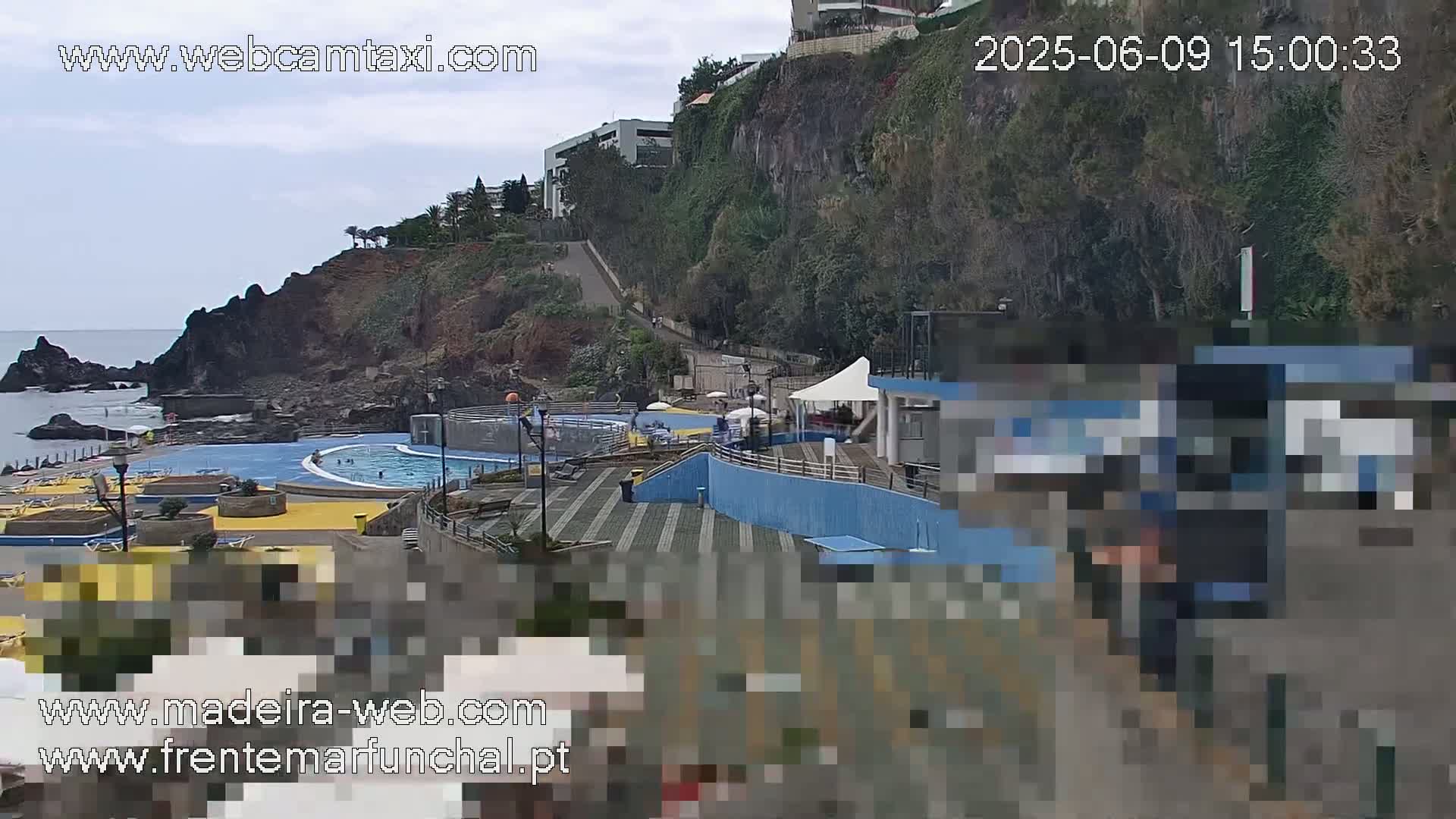 An outdoor scene shows an oceanside pool and patio area situated below a cliffside with buildings and vegetation, under an overcast sky.