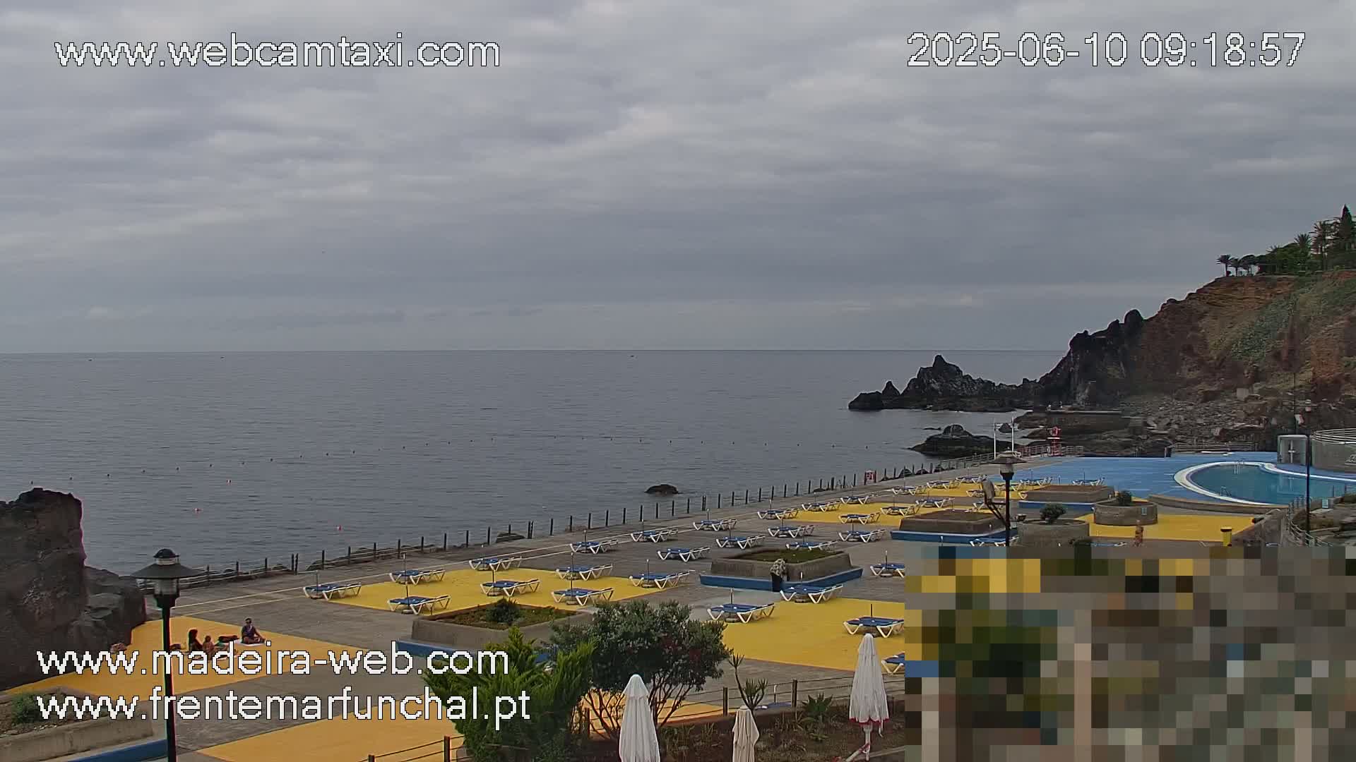An outdoor pool and deck area with numerous lounge chairs are situated on a rocky coastline overlooking a calm ocean under an overcast sky.